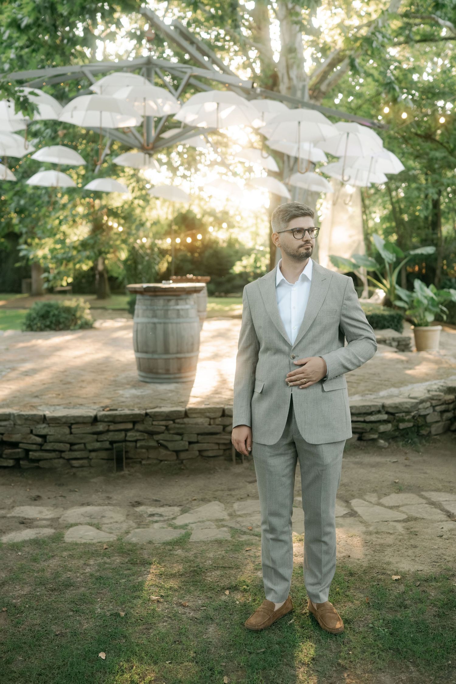 A person in a light gray suit holds a white umbrella while standing on grass near stone walls at sunset.