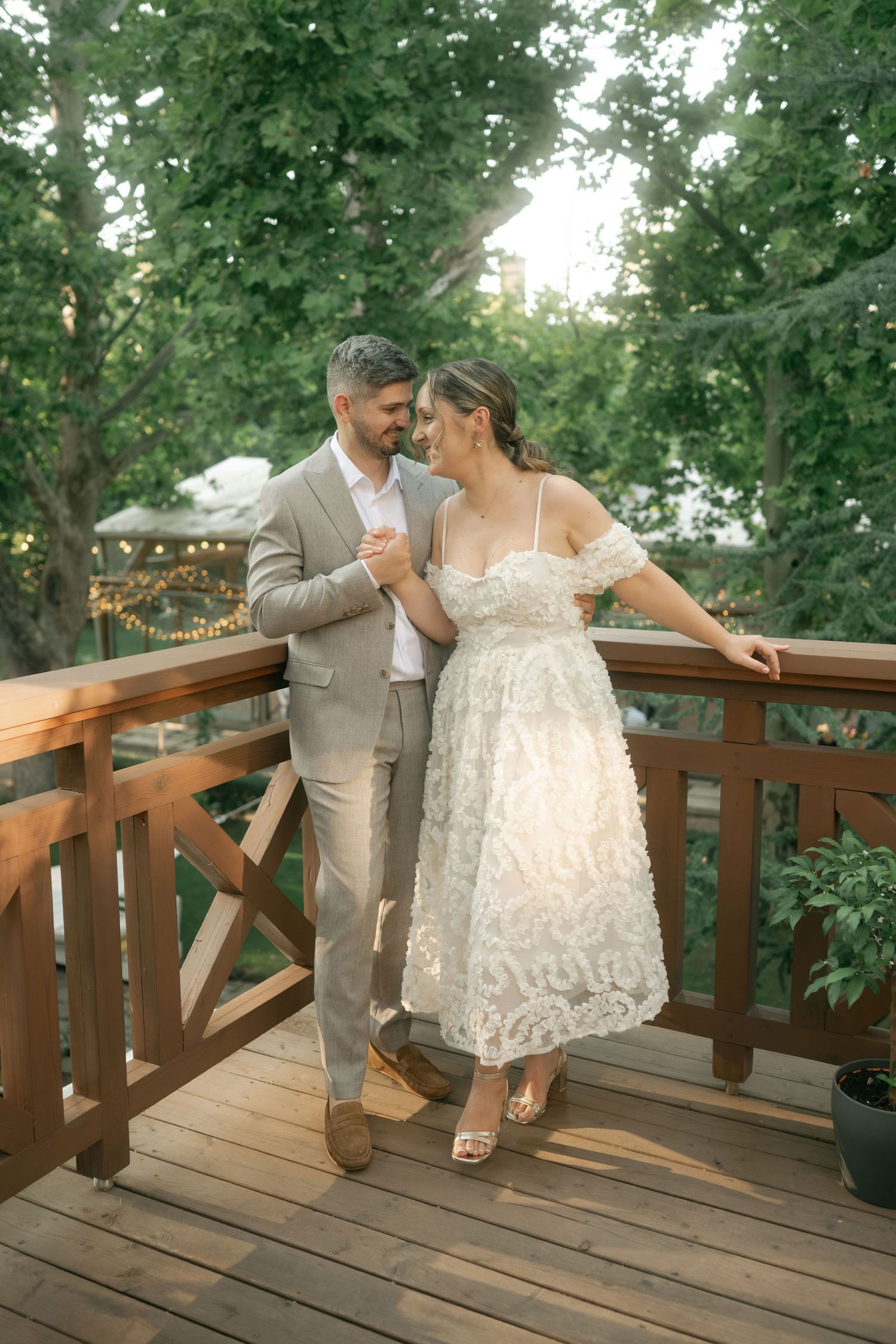 A couple in wedding attire dances playfully on a wooden deck surrounded by green trees.