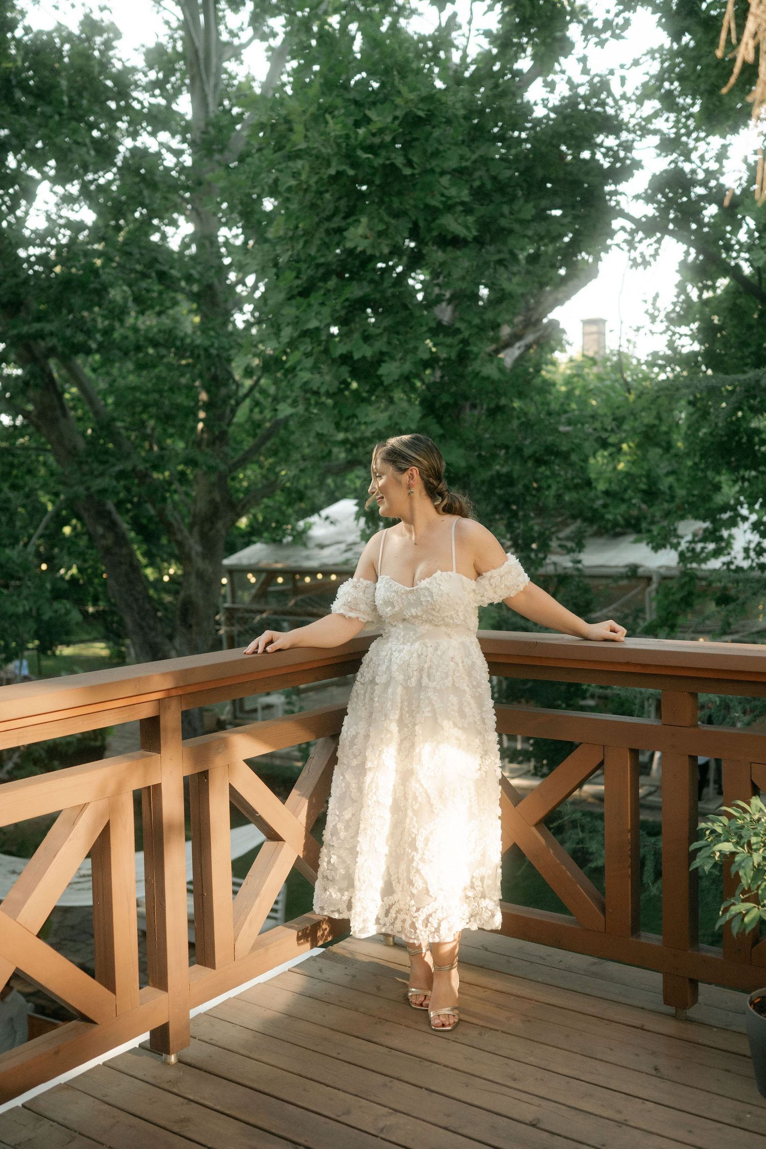 A woman in a flowing white dress poses gracefully on a wooden deck surrounded by lush green trees.