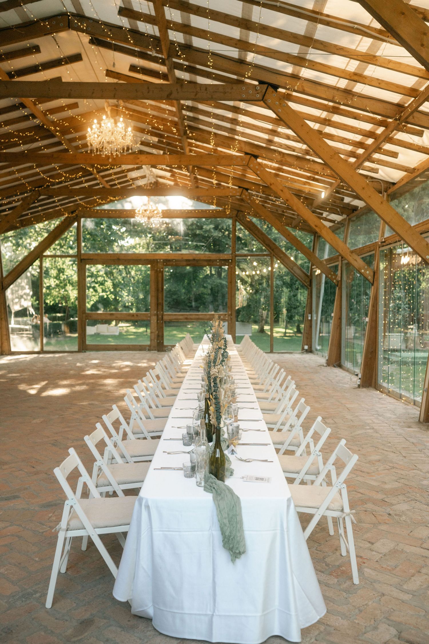 Rustic wooden barn venue set up for dining with white tables, chairs and elegant chandeliers hanging from exposed beams.