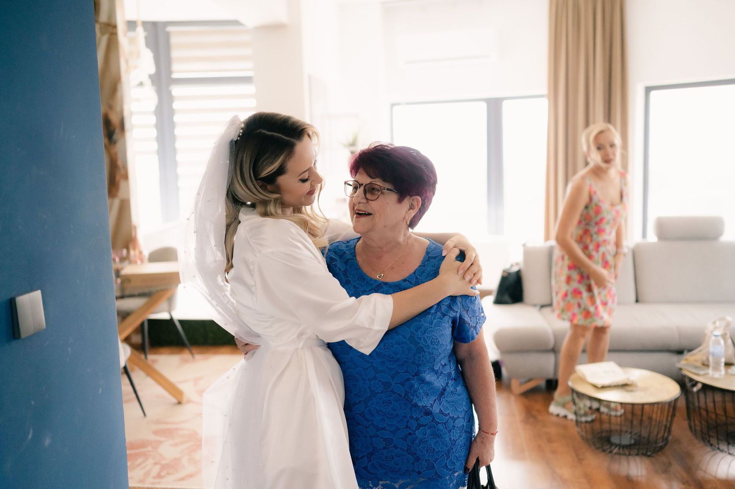 Two people share an emotional embrace in a sunlit living room while another person stands in the background.
