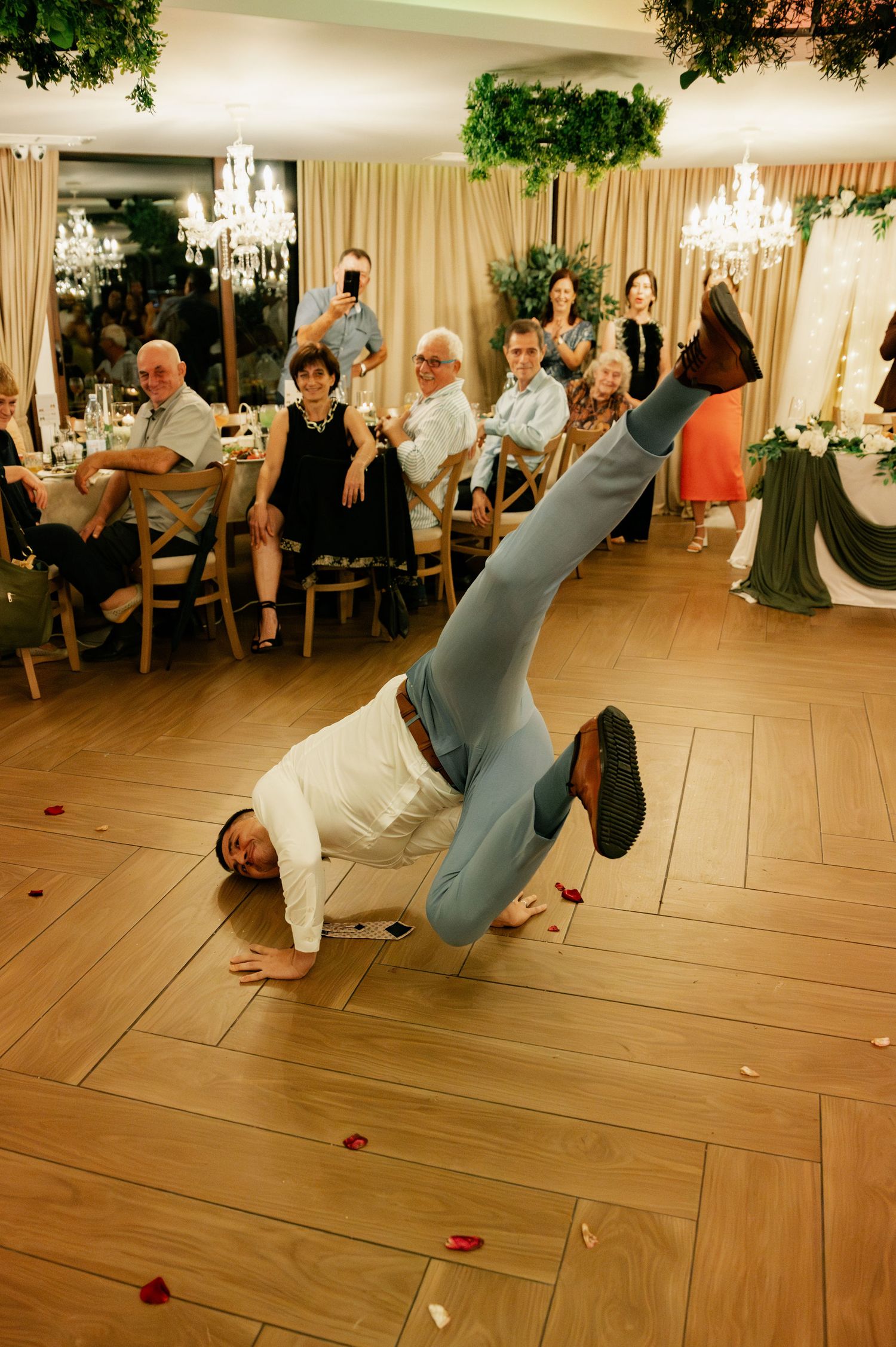 Wedding guests perform an energetic breakdancing routine on a wooden dance floor at an elegant reception venue.