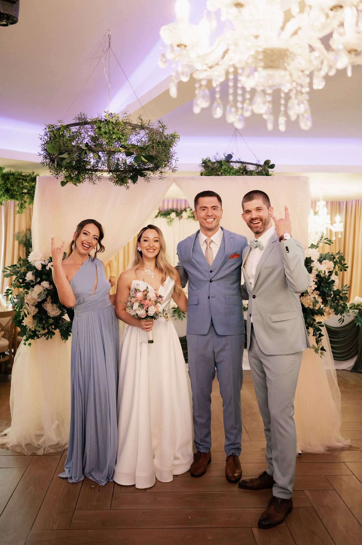 Wedding party in gray and white attire poses under elegant chandeliers in a reception venue.