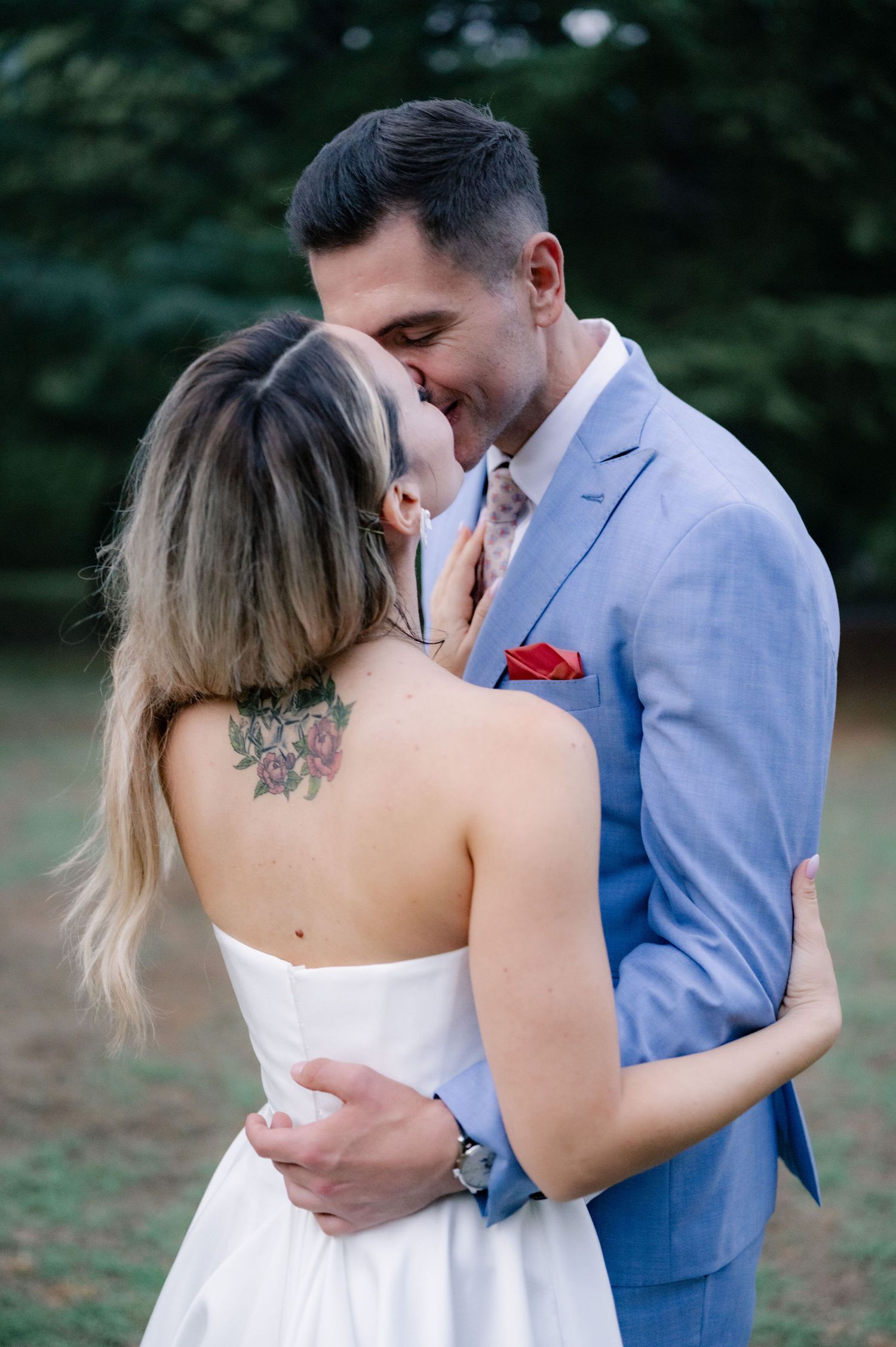 A couple in formal attire share an intimate embrace outdoors, with the woman wearing white and the man in a blue suit.