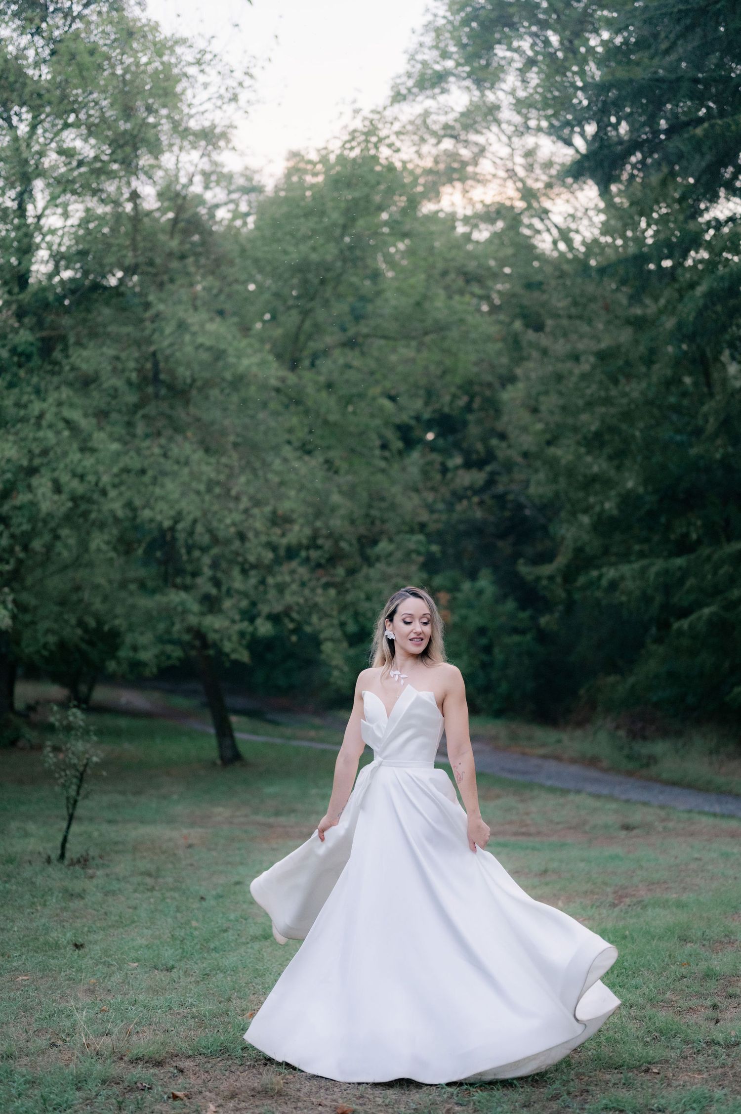 A person in a flowing white wedding dress twirls gracefully in a natural outdoor setting among trees and grass.