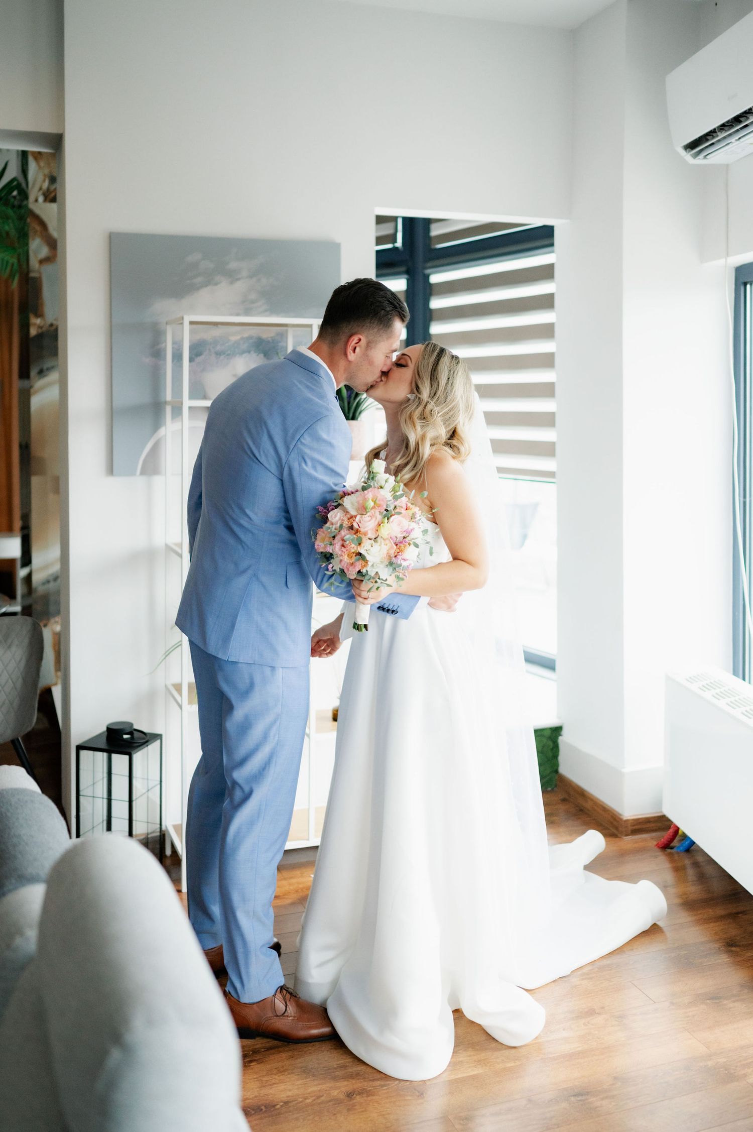 A couple in wedding attire share an intimate moment in a bright modern room with hardwood floors and window light.