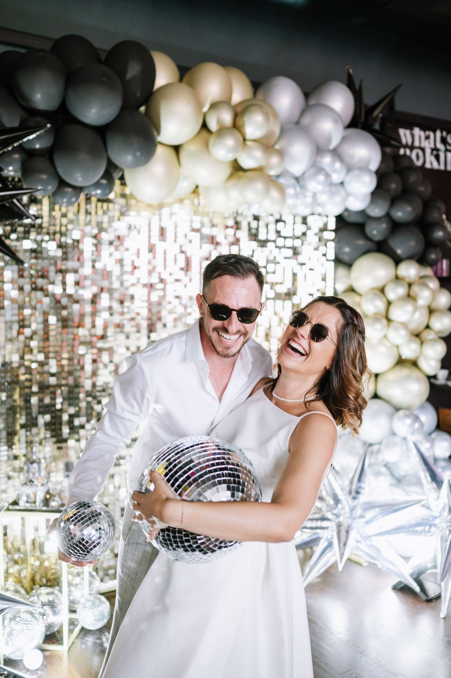 A couple dances and laughs together under a ceiling decorated with silver disco balls and white balloons.