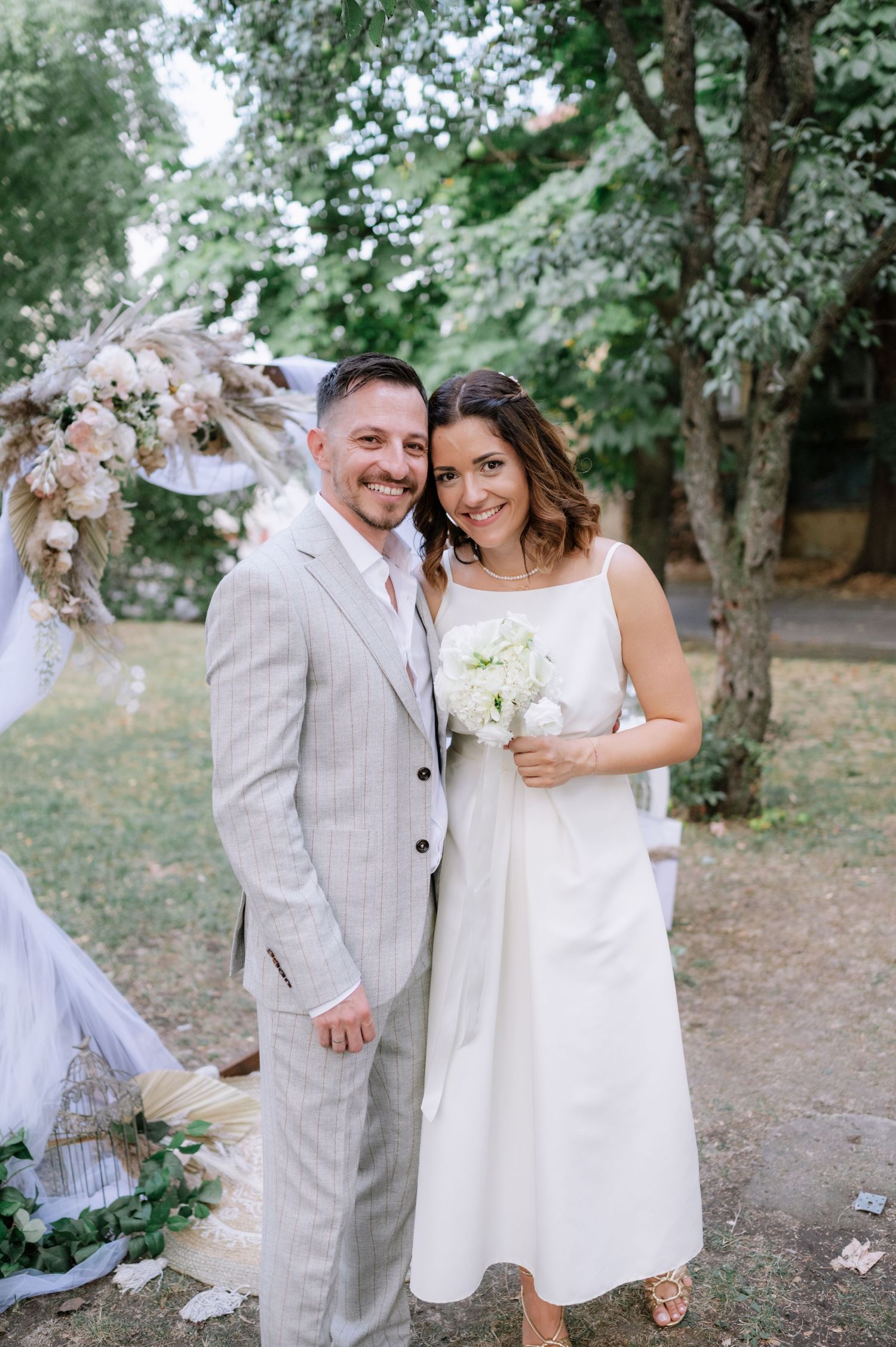 A couple shares romantic moments in a wooded outdoor setting wearing light-colored wedding attire and holding a bouquet.