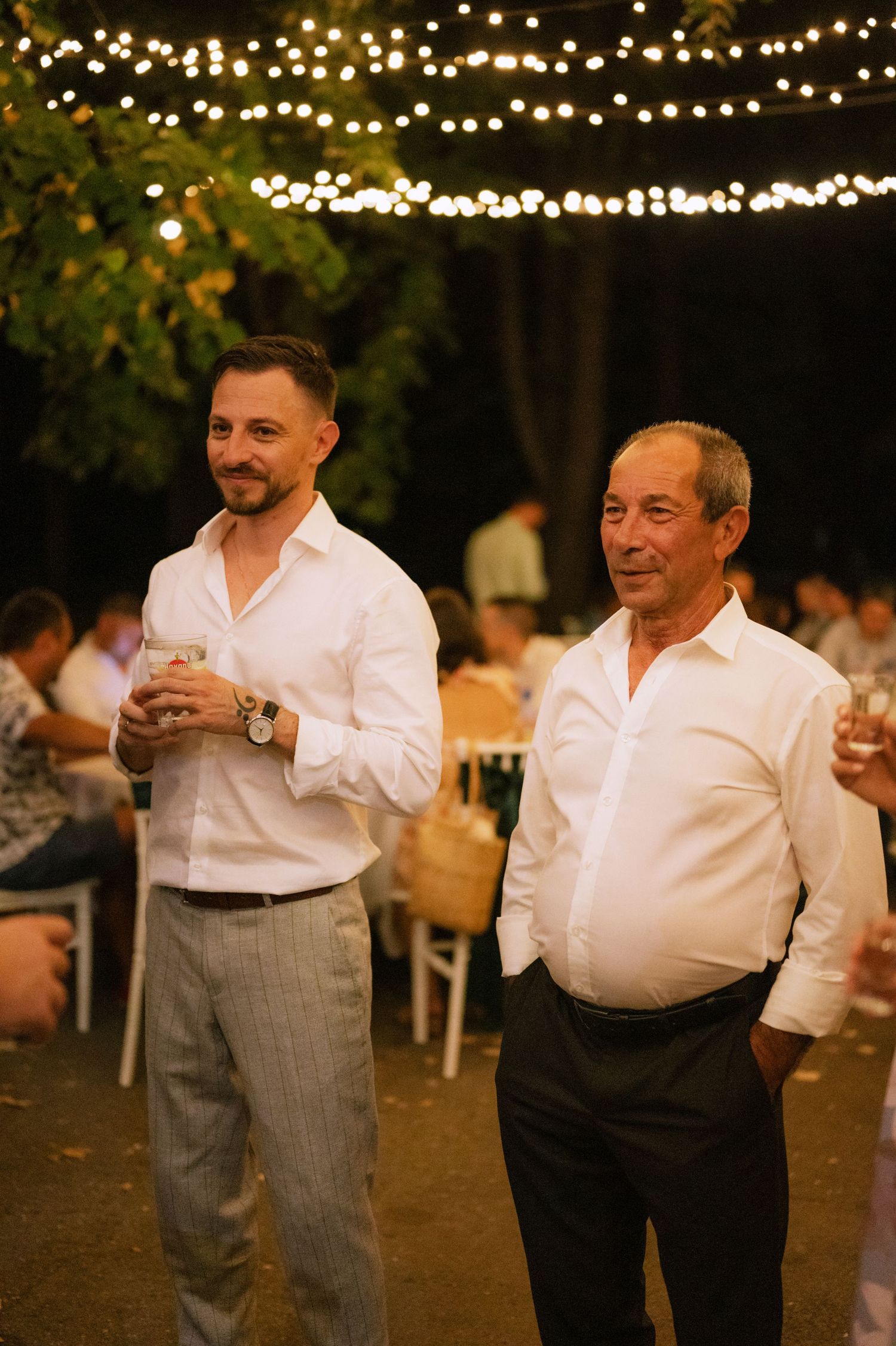 Two men in white shirts and grey pants dancing together at an outdoor evening event with string lights overhead.