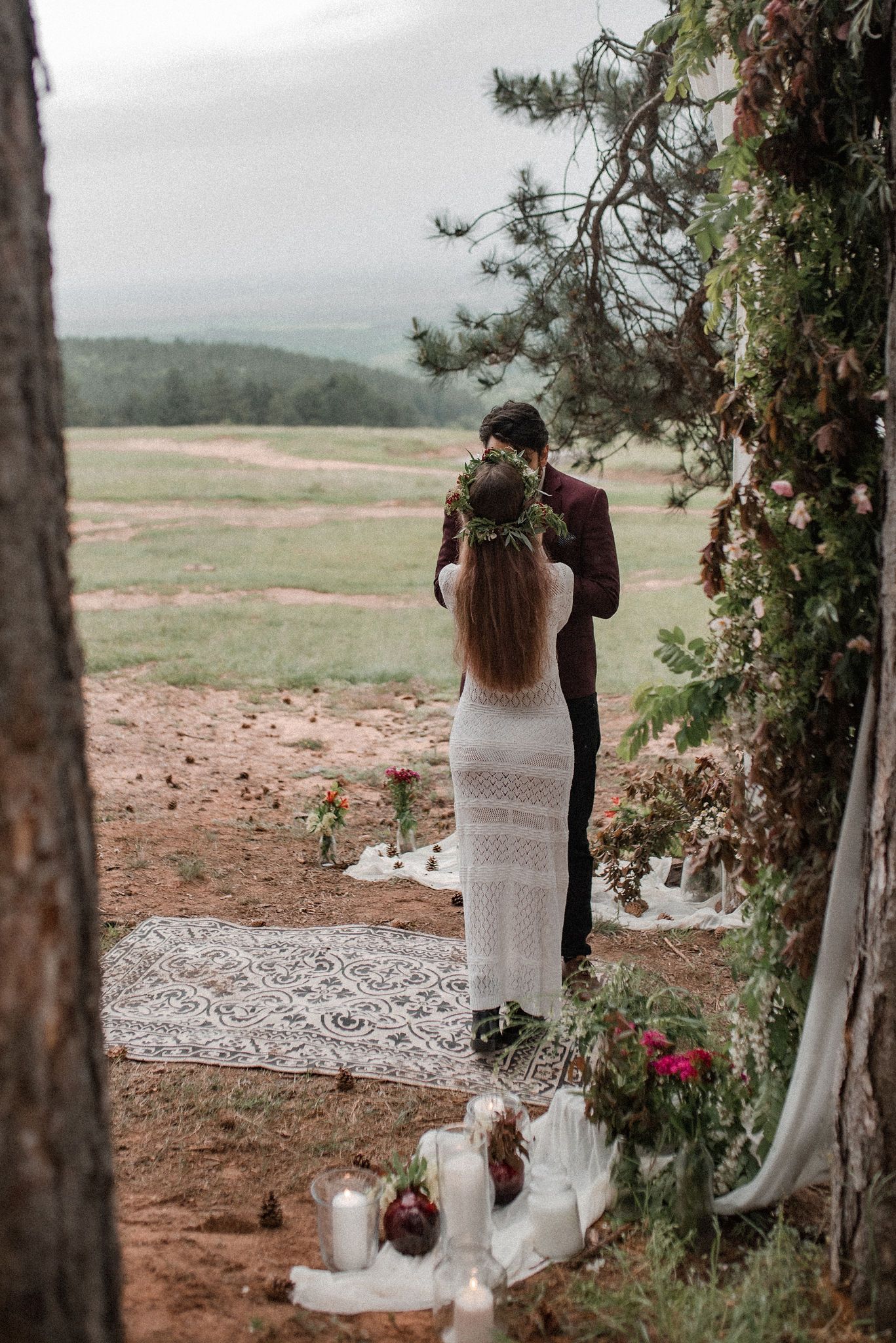 Rustic outdoor wedding ceremony setup with evergreen trees and white fabric draped between trunks overlooking a scenic landscape.