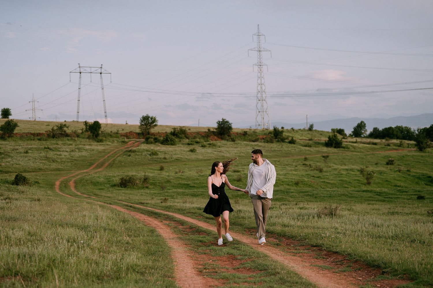 Two people walk together along a winding dirt path through a green prairie landscape with power lines in the distance.