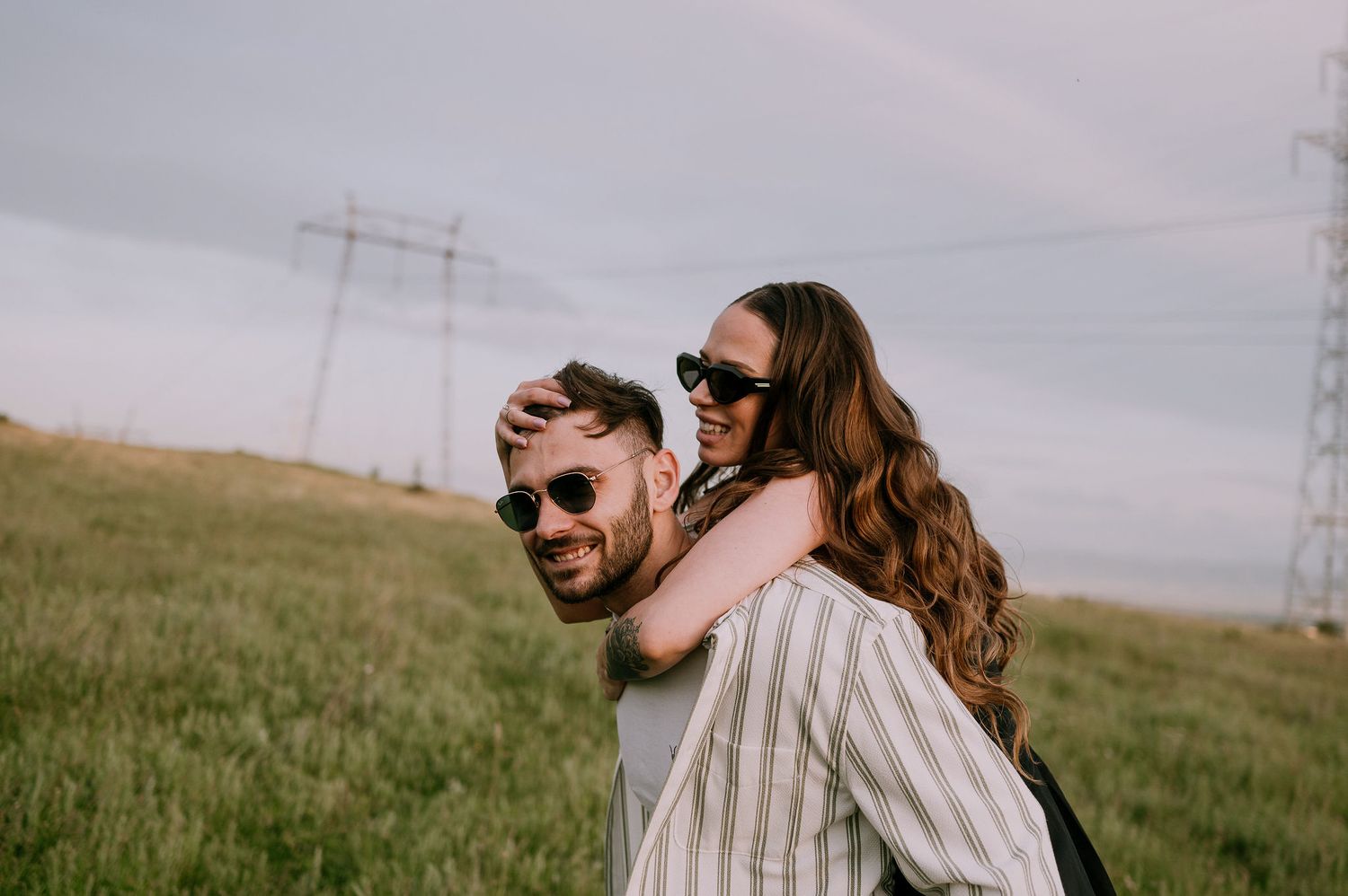 A couple embraces and laughs together in a grassy field with power lines visible in the background on an overcast day.