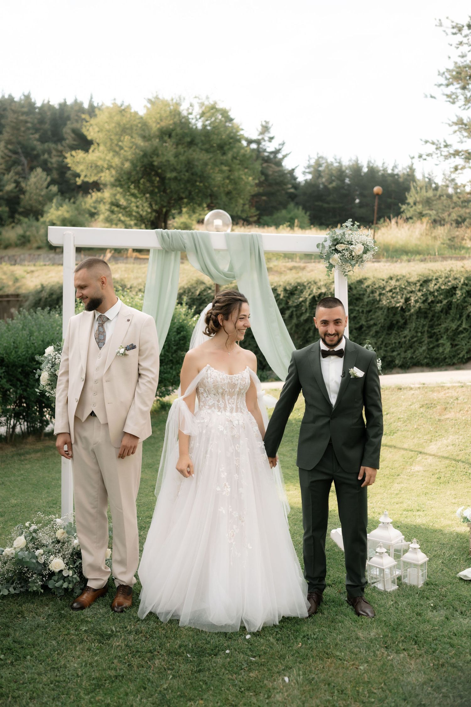 Wedding party members walk together on a lush green lawn with white outdoor ceremony decor visible in summer.