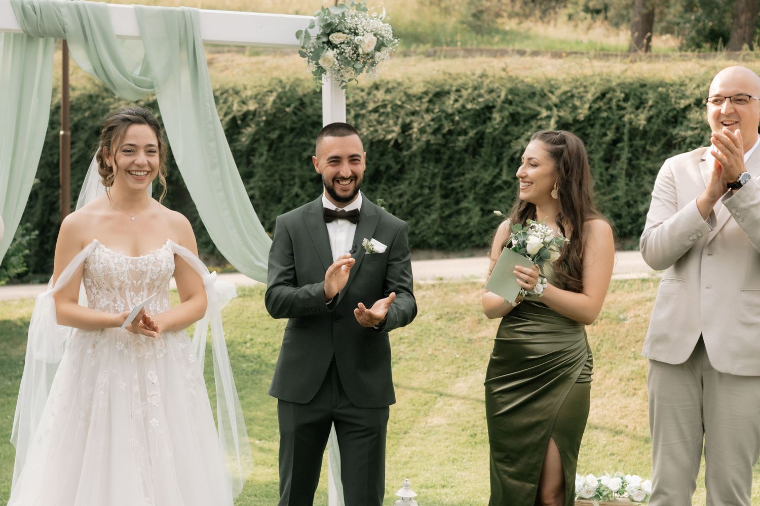 Wedding party celebrating outdoors with joyful reactions under white fabric draping and natural lighting.