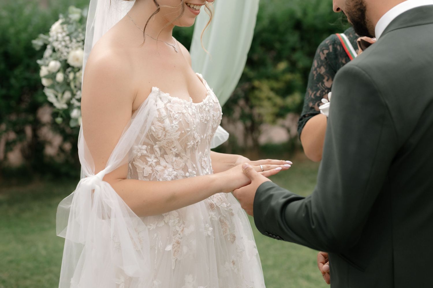 Couple holding hands during outdoor wedding ceremony with lush greenery and white floral decor in background.