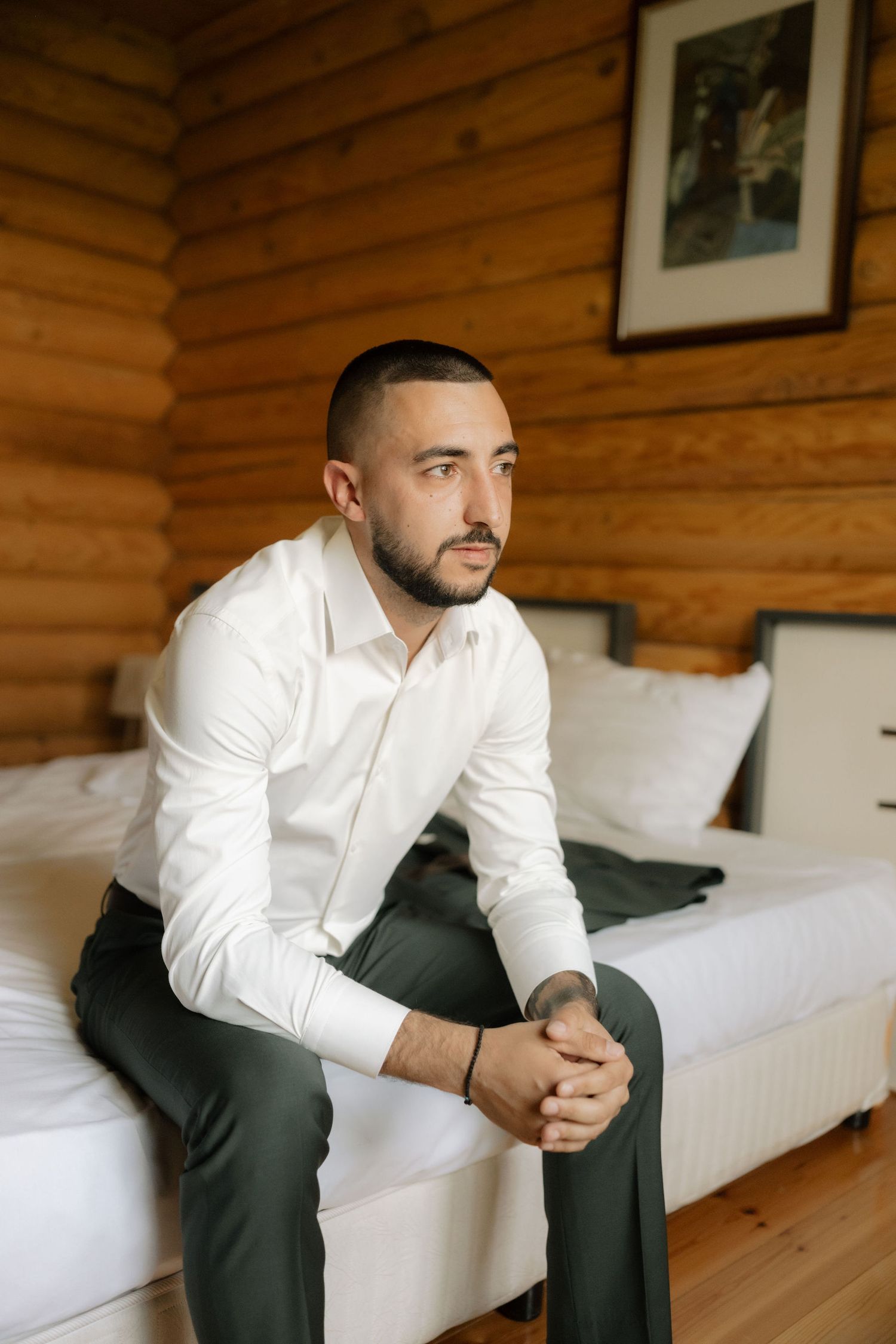 A person in a white dress shirt and dark pants sits thoughtfully on a white couch in a wood-paneled room.