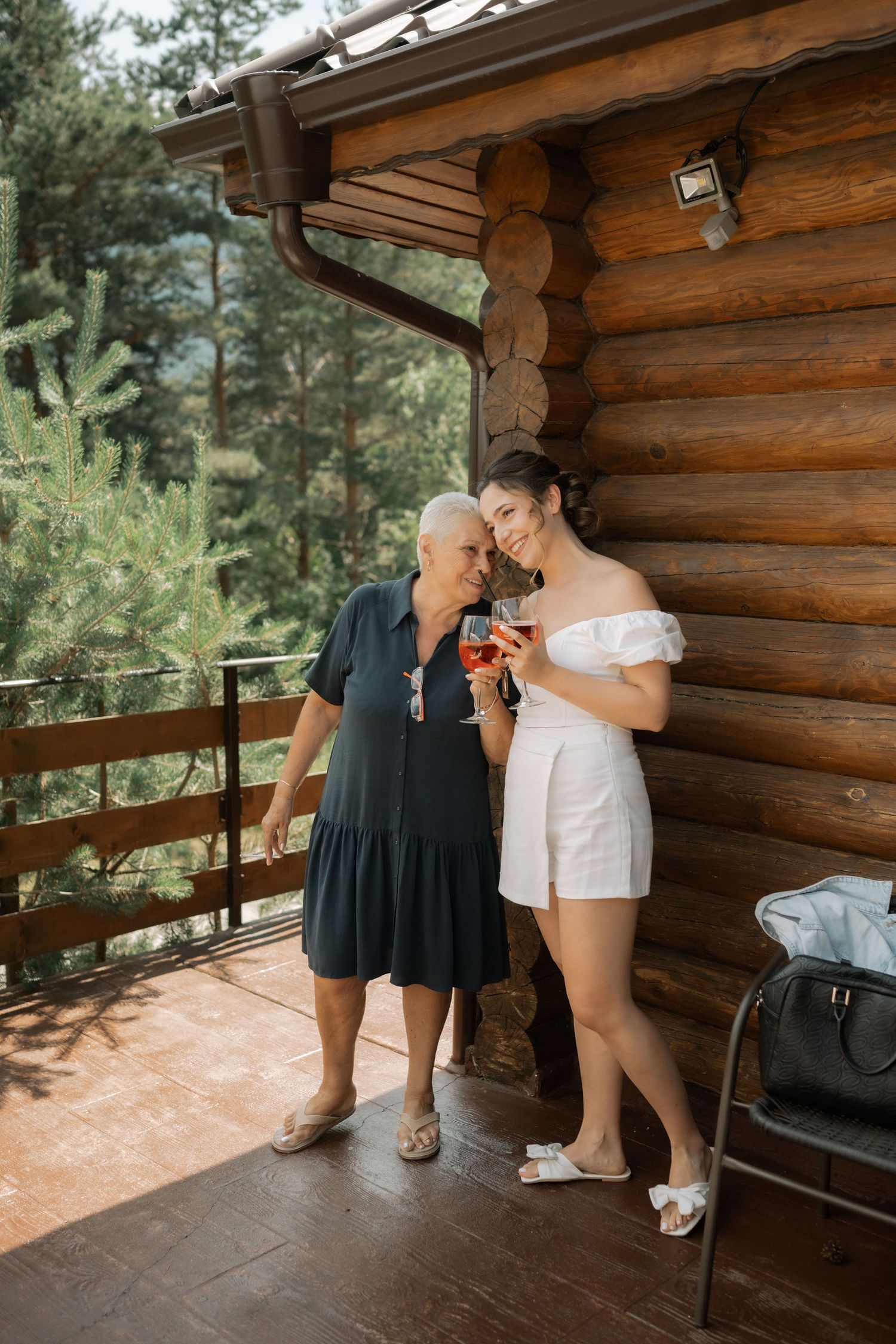 Two people share an intimate moment on the porch of a rustic log cabin surrounded by pine trees.