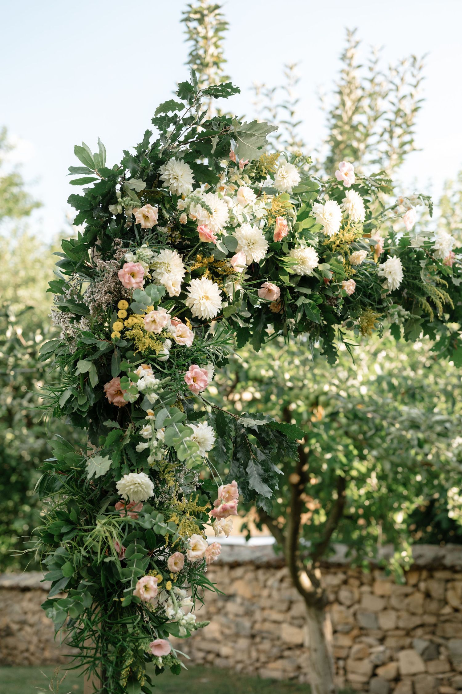 Elegant floral arch with white and pink blooms adorning a rustic stone wall in a garden setting.