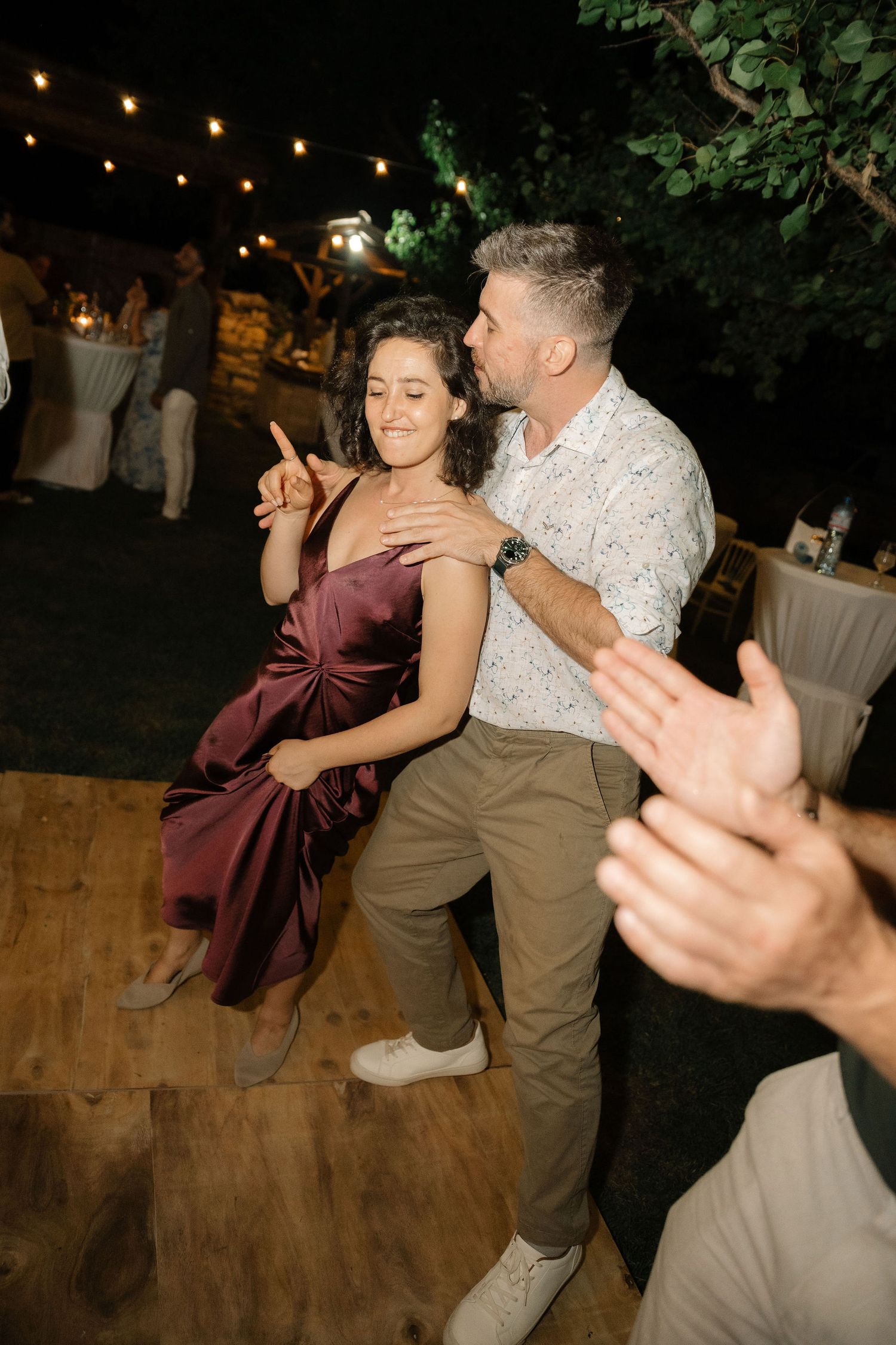 Dancers twirling and having fun on a wooden dance floor at an outdoor evening reception with string lights overhead.