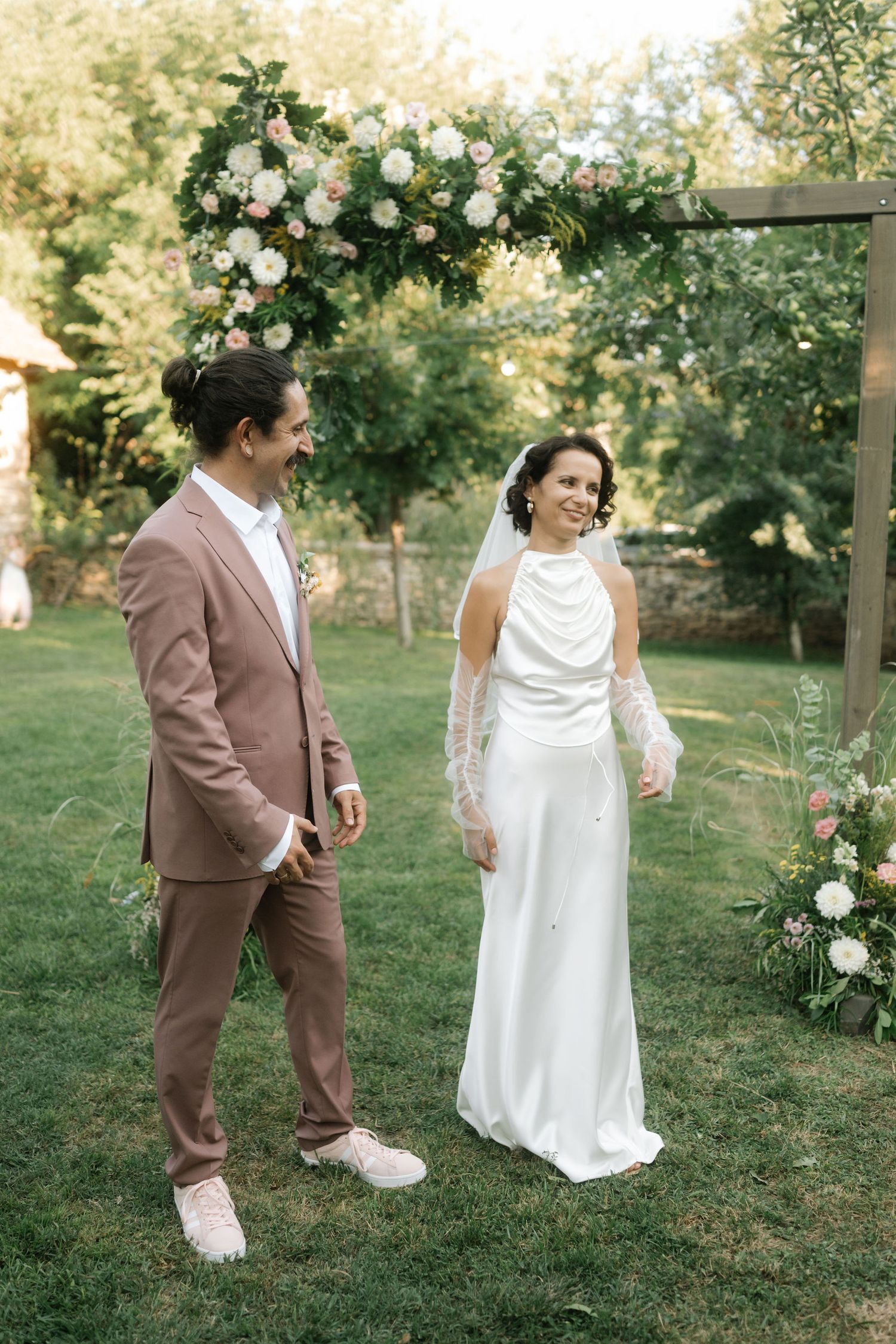 A couple in brown suit and white dress share candid moments in a garden setting with blooming roses.