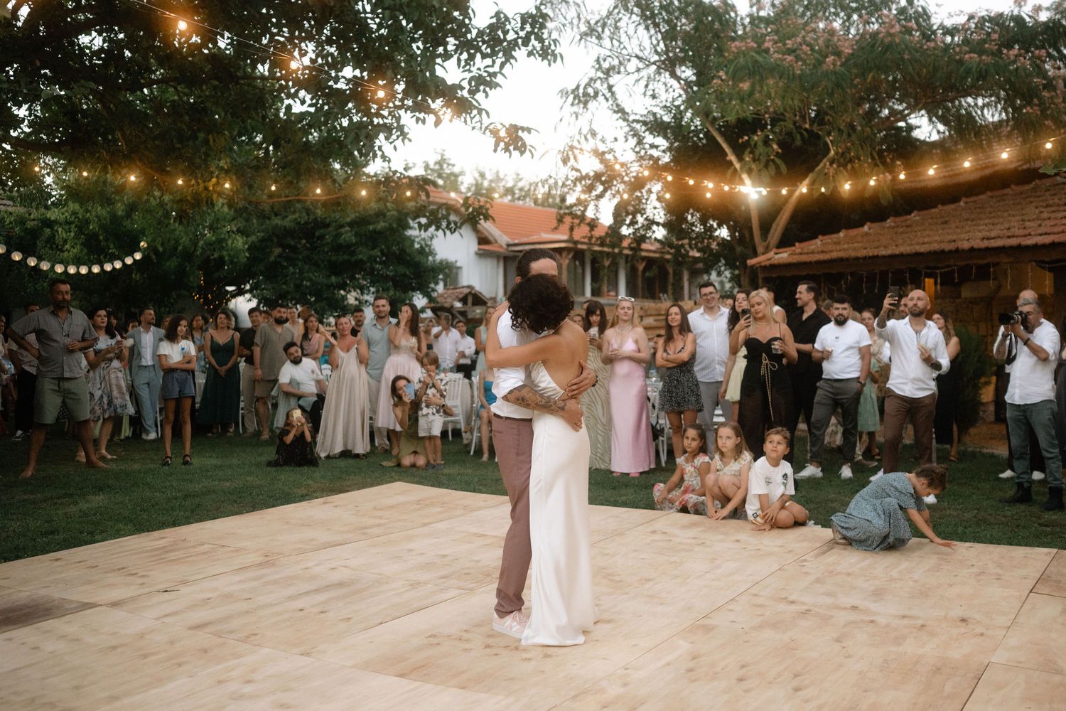 Wedding guests gather around a wooden dance floor as a couple dances beneath string lights at an outdoor reception.