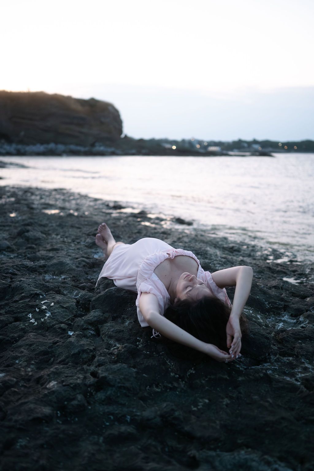 A person in light clothing rests on a black sand beach with coastal cliffs visible in the background at sunset.