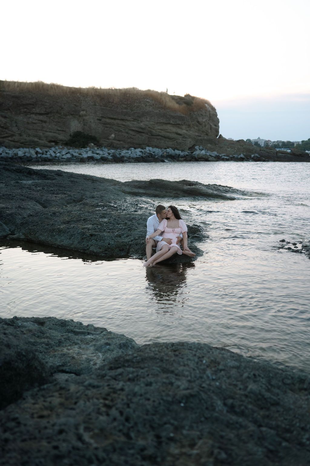 A couple shares an intimate moment on dark volcanic rocks by the ocean at sunset.