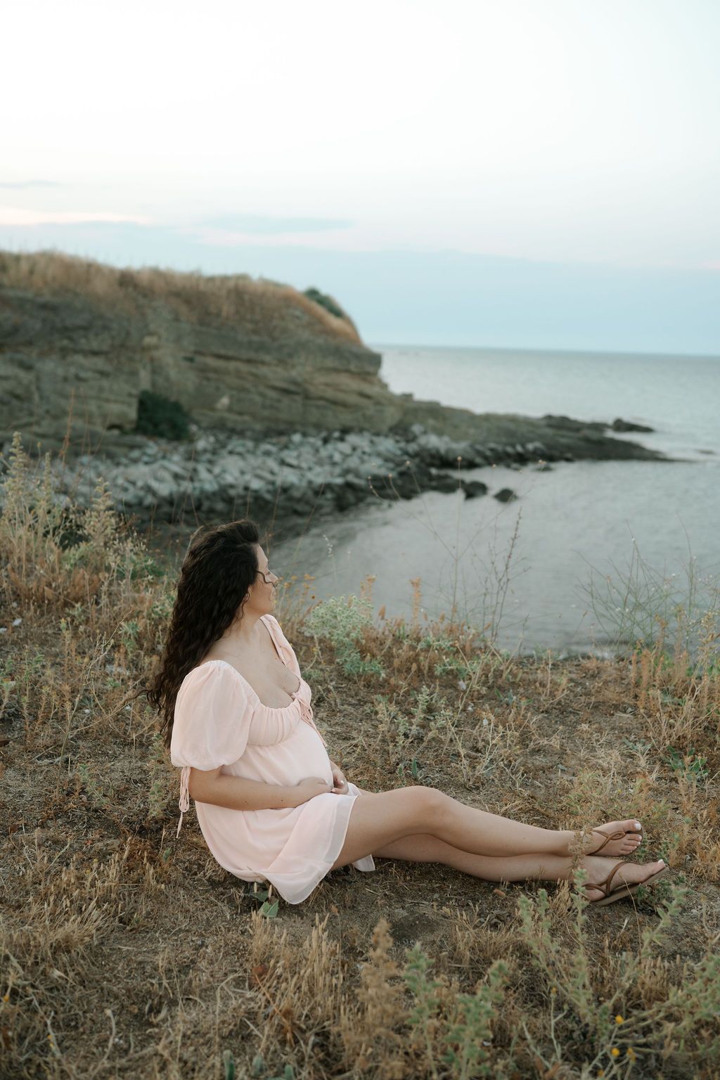 A figure in a pink dress reclines on rocky coastal terrain at sunset with dramatic cliffs and ocean in the background.