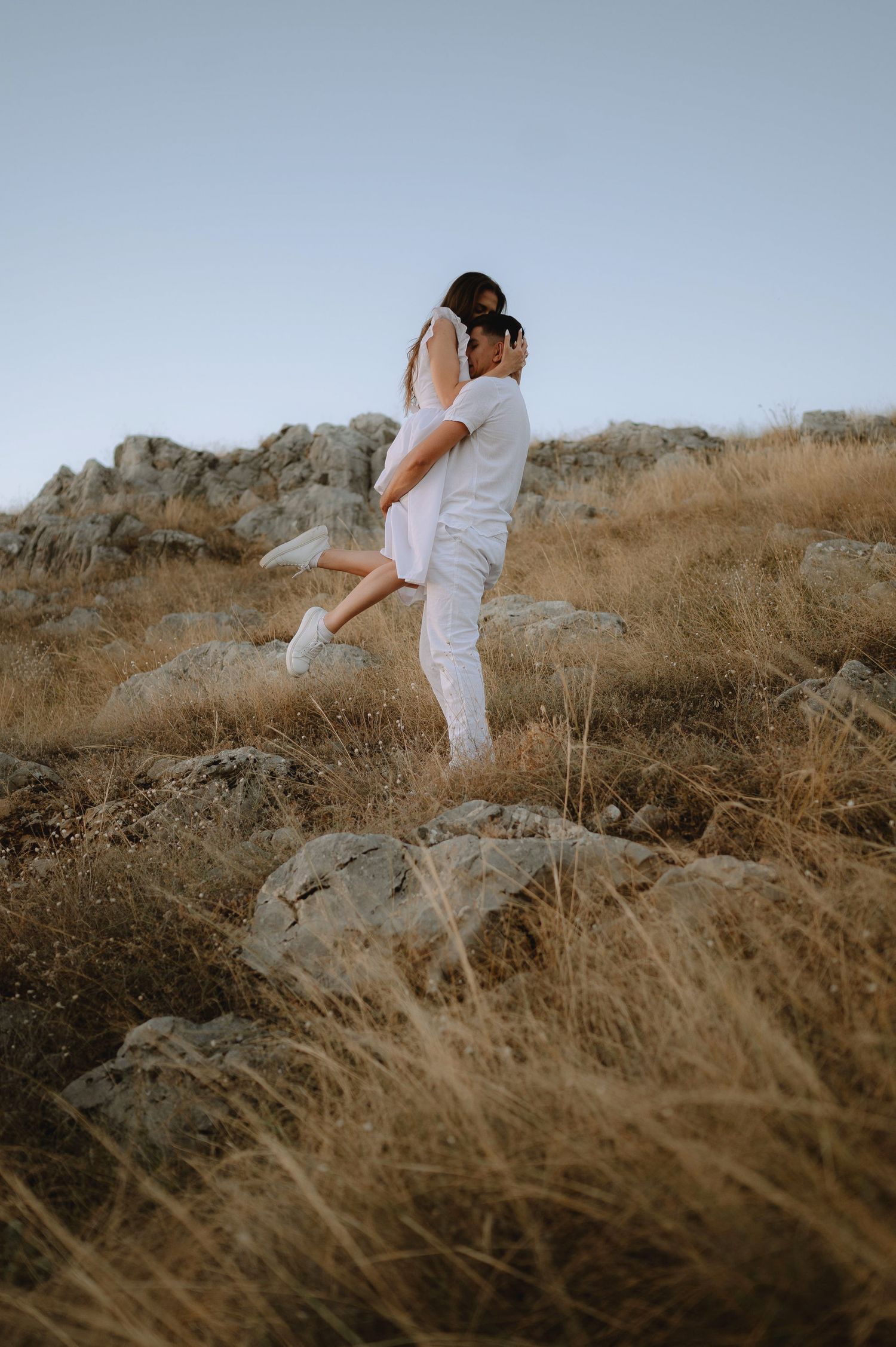 A couple in white outfits embraces on a hillside with golden dried grass and rocks at sunset.