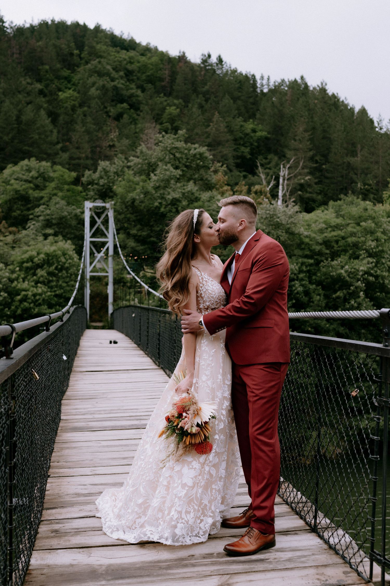 A couple in wedding attire shares romantic moments on a rustic suspension footbridge surrounded by lush forest scenery.