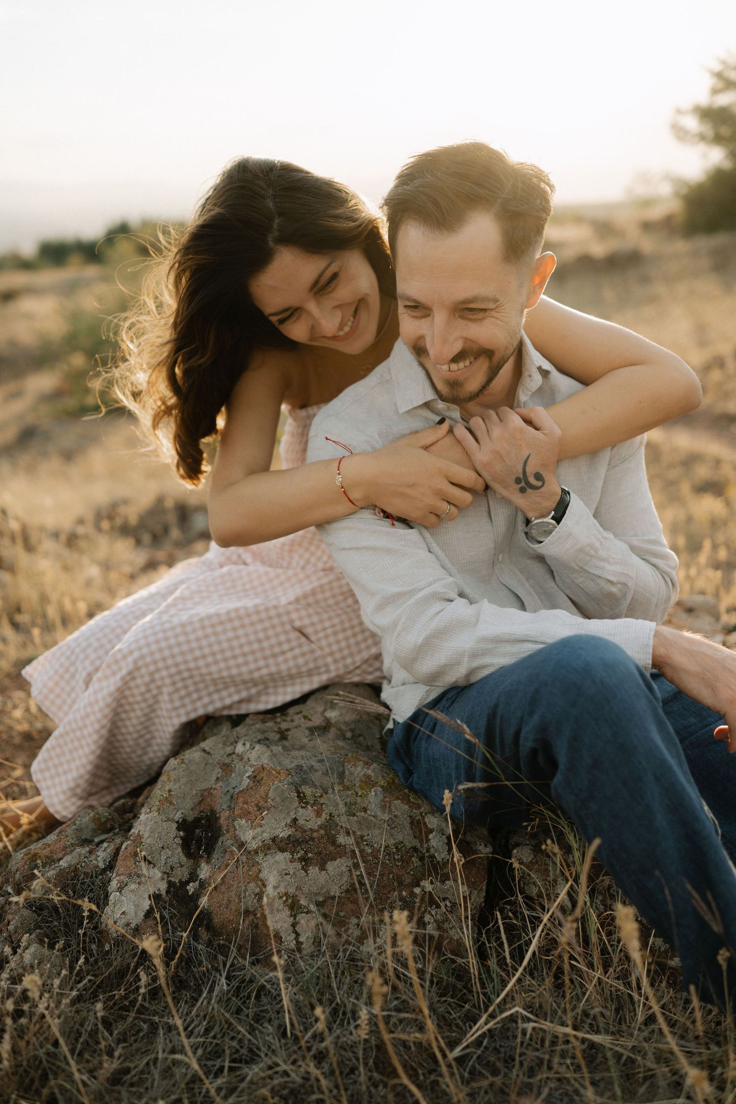 A couple shares an intimate moment while sitting on a rock in a golden field during sunset.