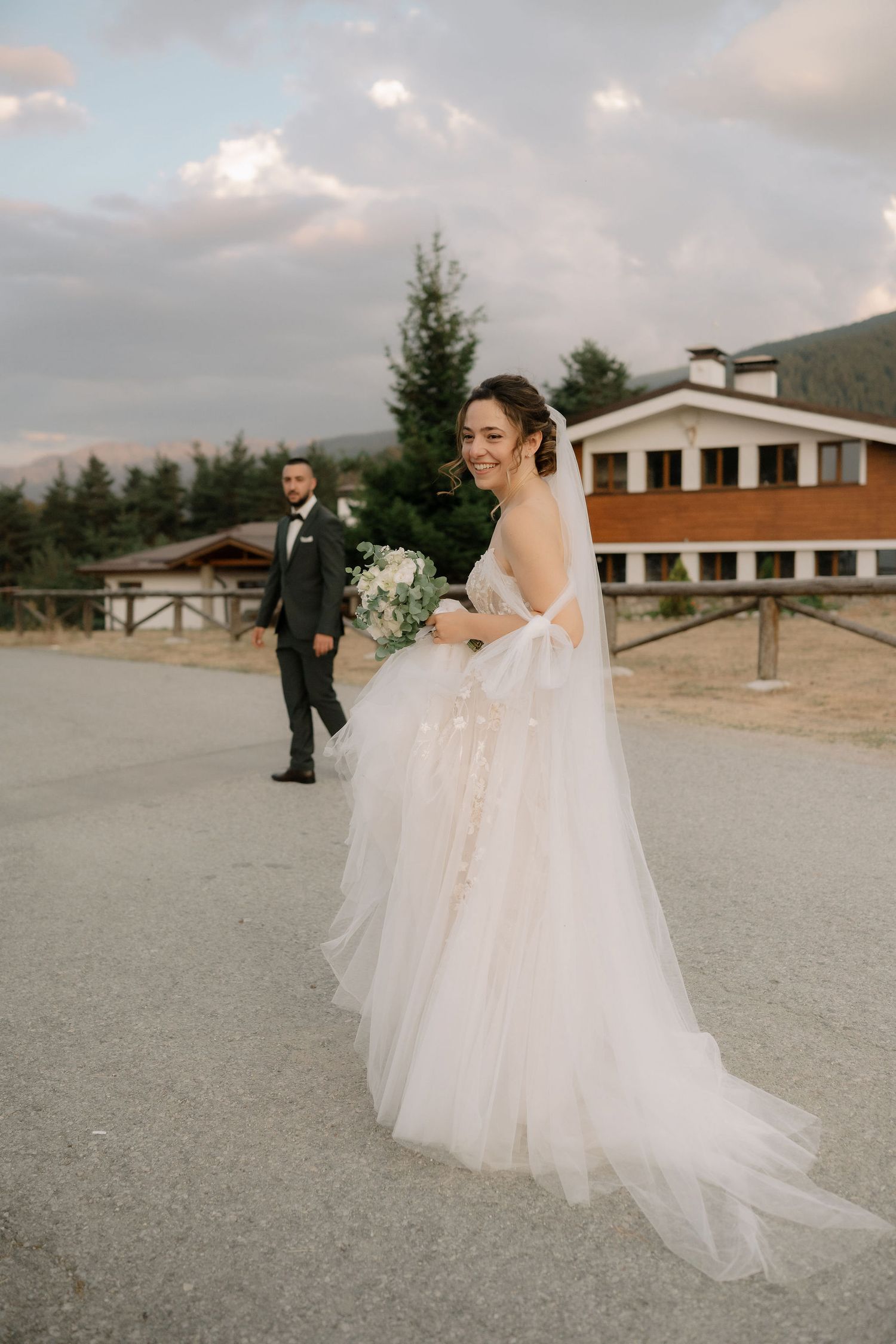 A wedding couple walks together on a gravel path with mountains and buildings in the background.