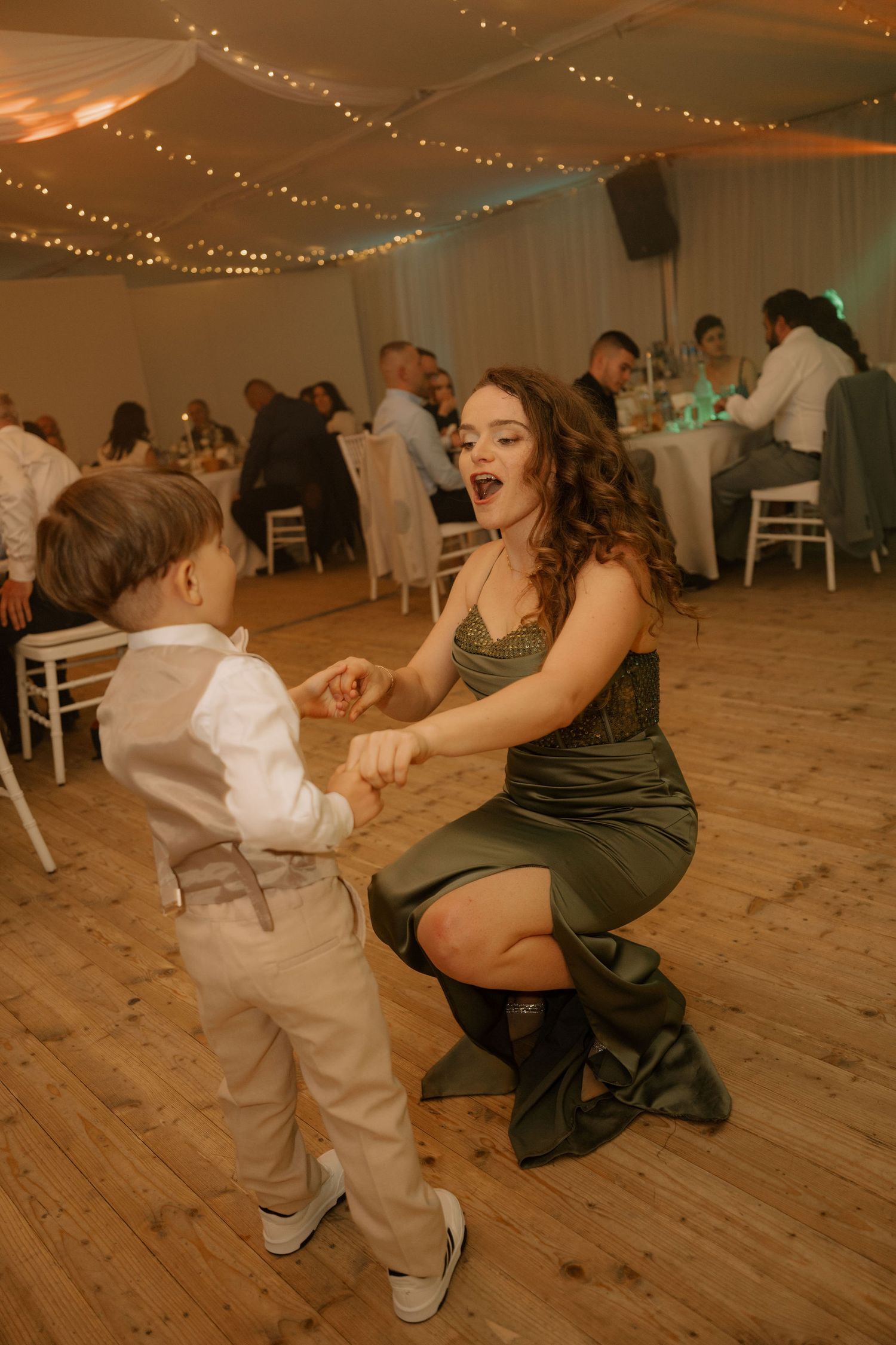 Woman in green dress dancing joyfully with young boy in suspenders at elegant wedding reception.
