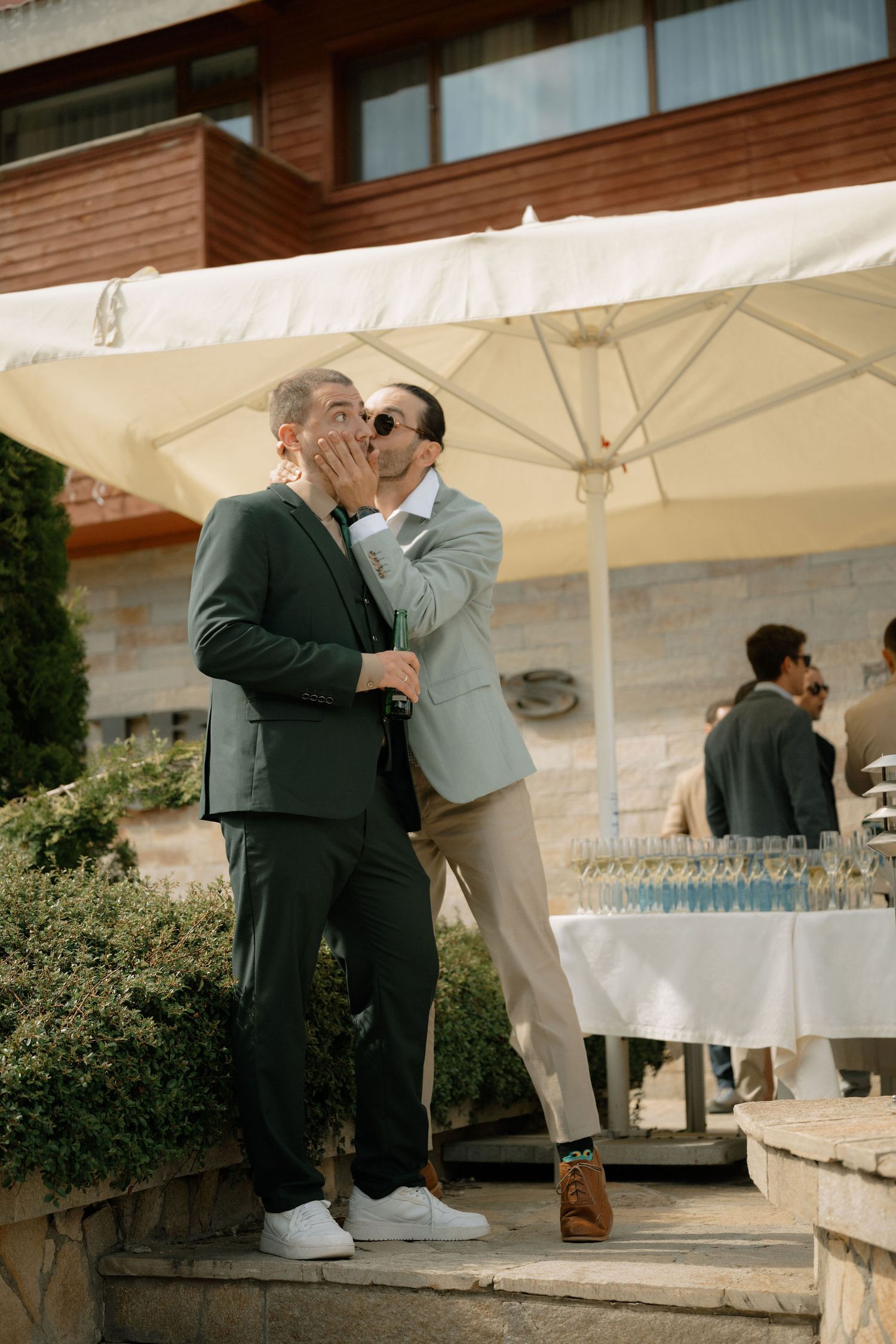 Two grooms in suits share a kiss outdoors at their wedding reception near a champagne display.