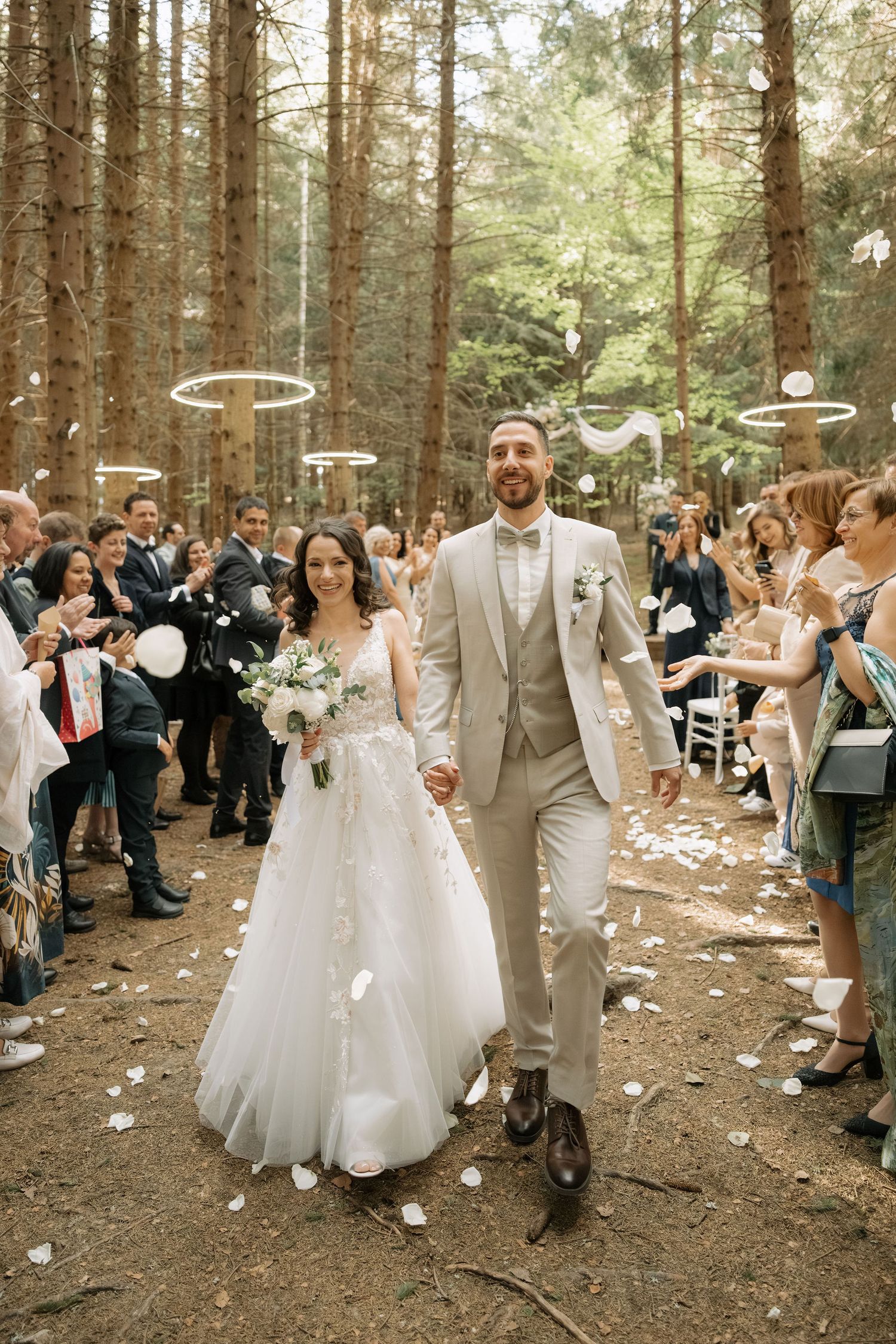 Bride and groom walk down forest aisle as guests toss white petals during outdoor woodland wedding ceremony.
