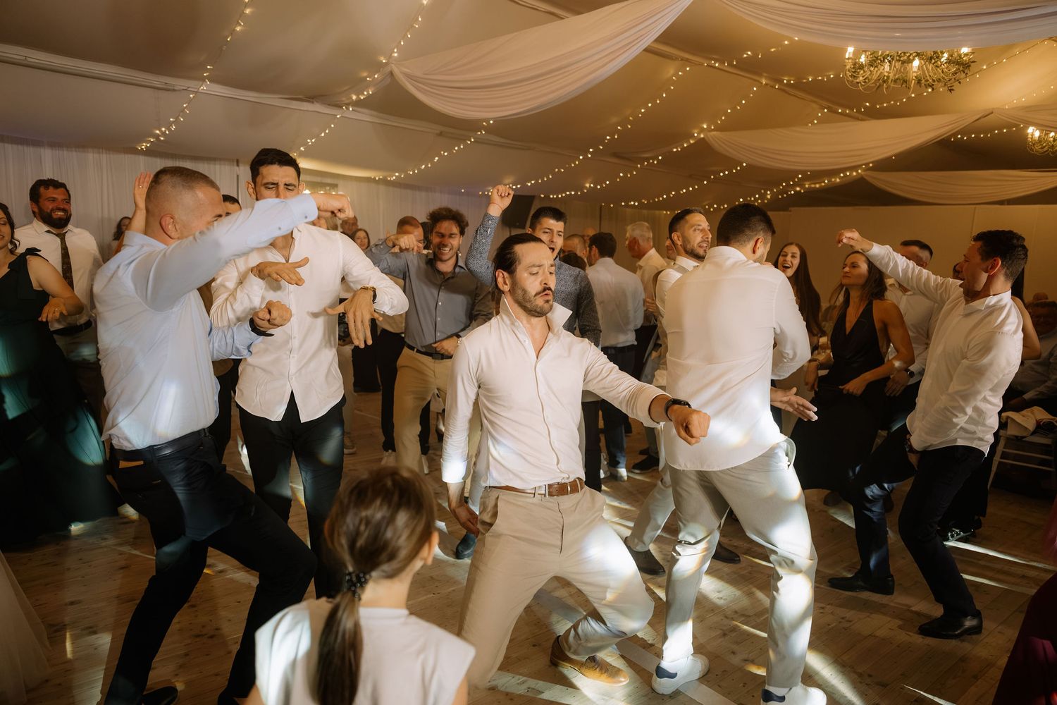 Guests dancing energetically at a wedding reception in a decorated ballroom with string lights.