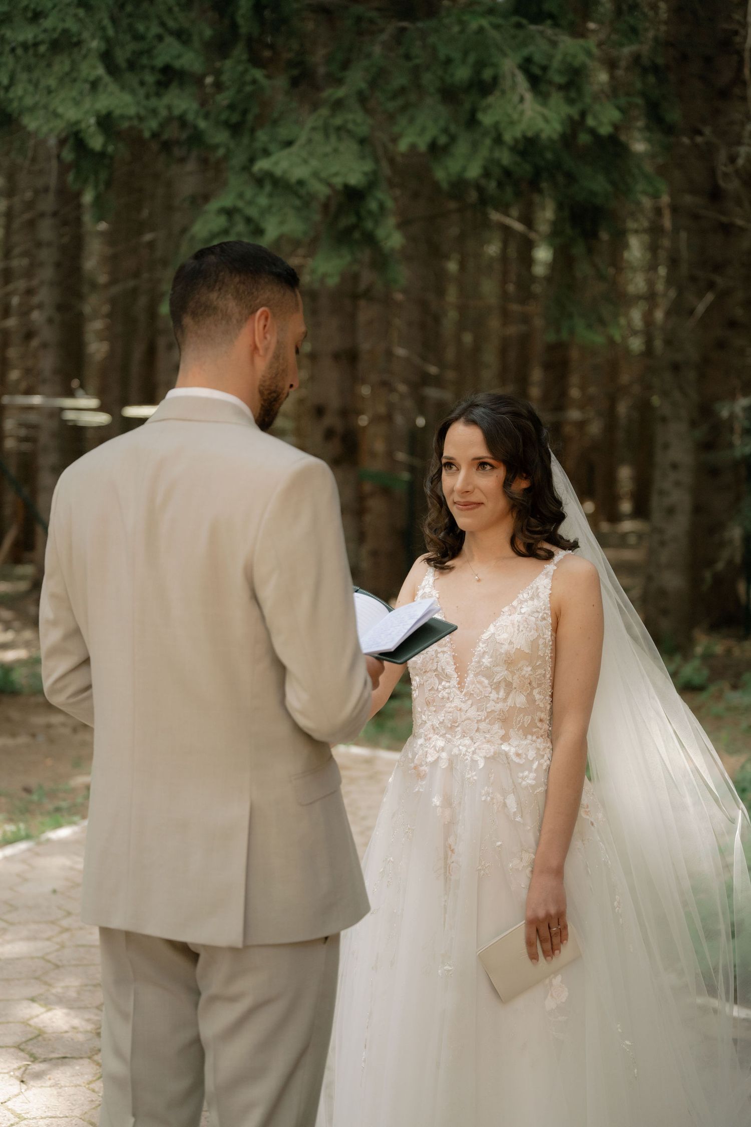 Bride in lace gown and veil exchanges vows with groom in beige suit during outdoor forest wedding ceremony.