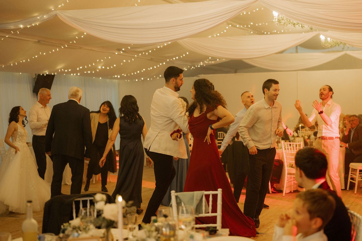 Guests dancing and celebrating at an elegant wedding reception in a decorated marquee with fairy lights.