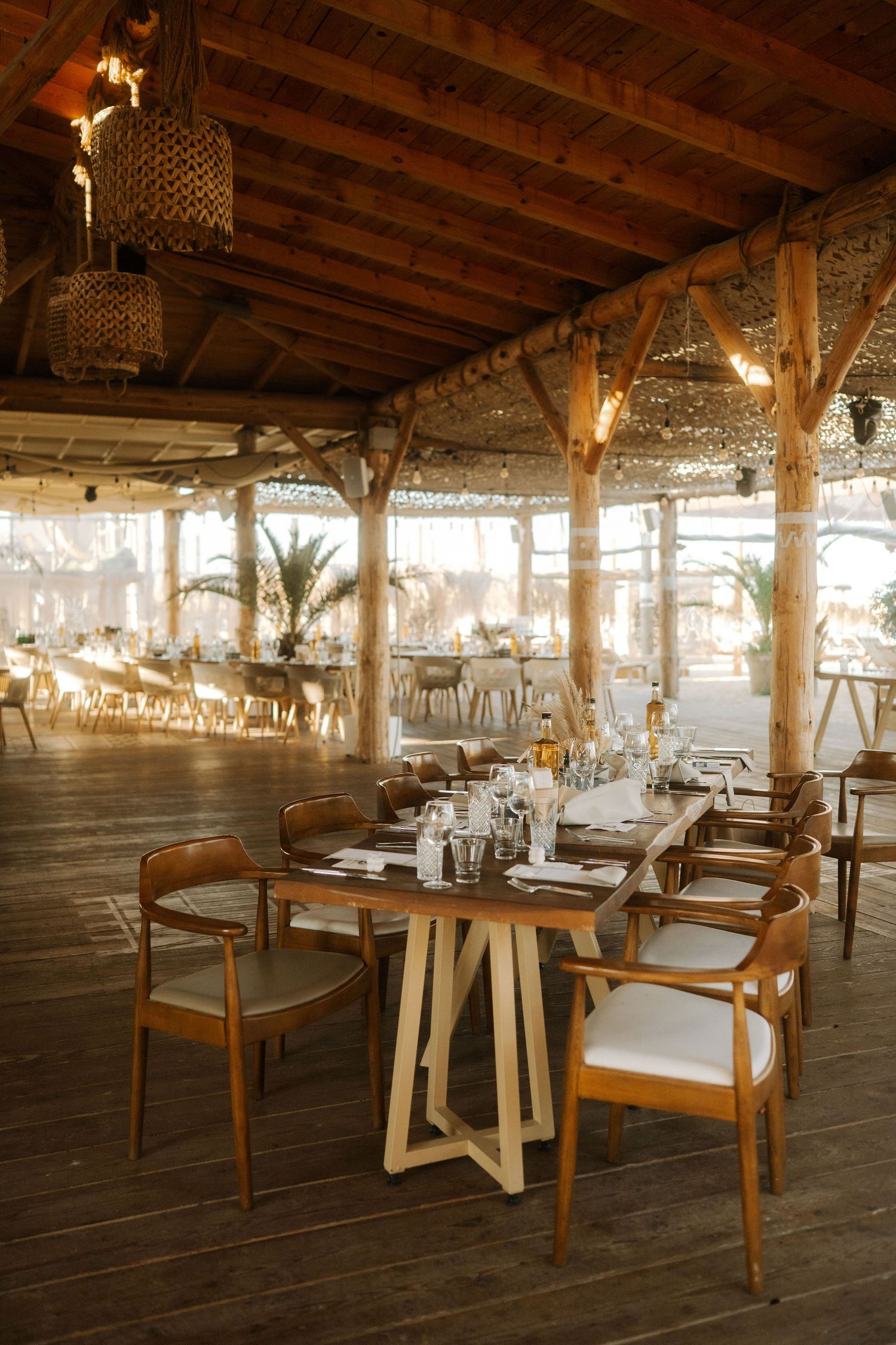 Rustic wooden event venue interior with elegant table settings, crystal chandeliers, and exposed beam ceiling.