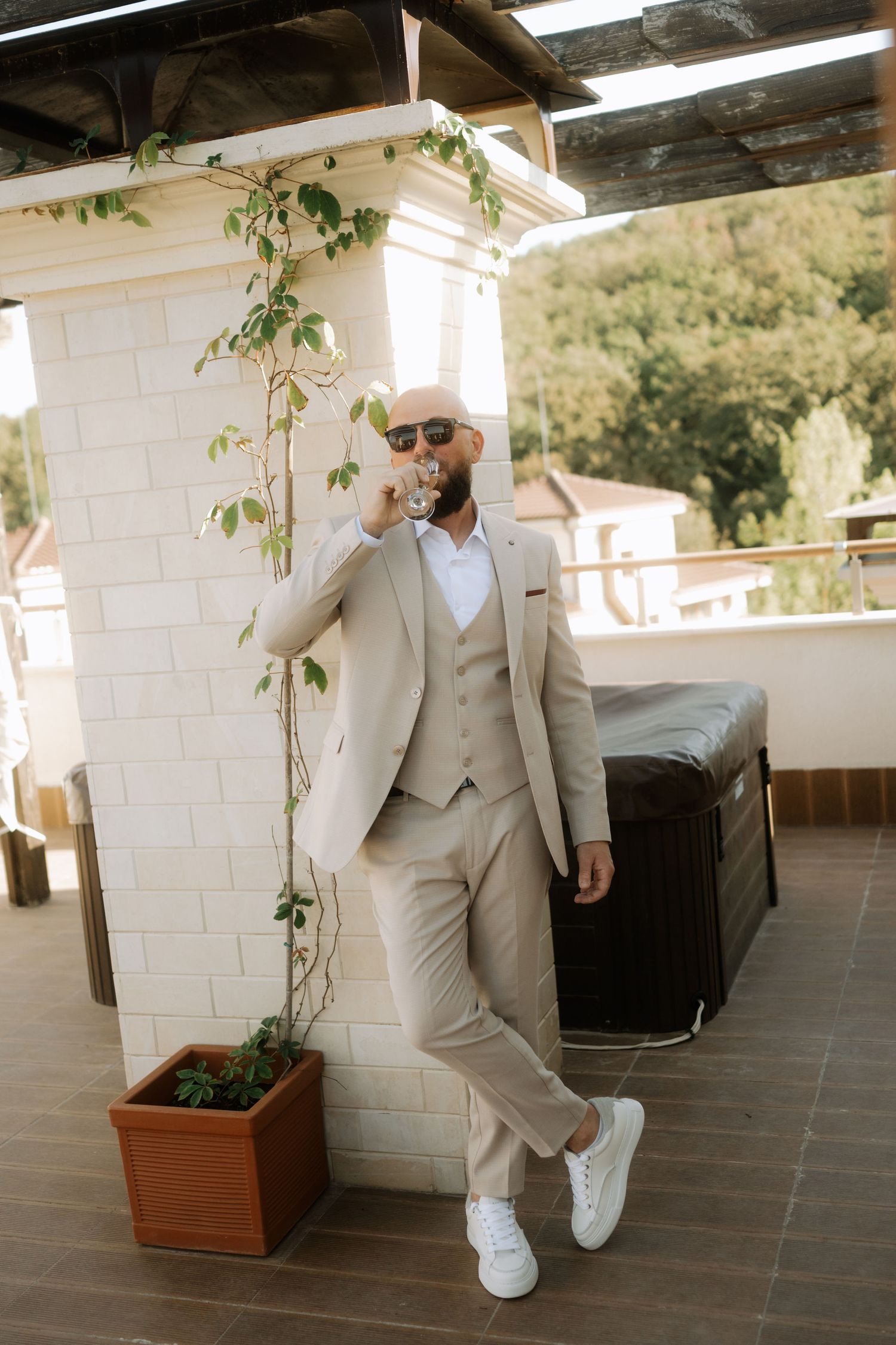 A figure in a light beige suit and white sneakers poses casually against a white wall in an outdoor setting.