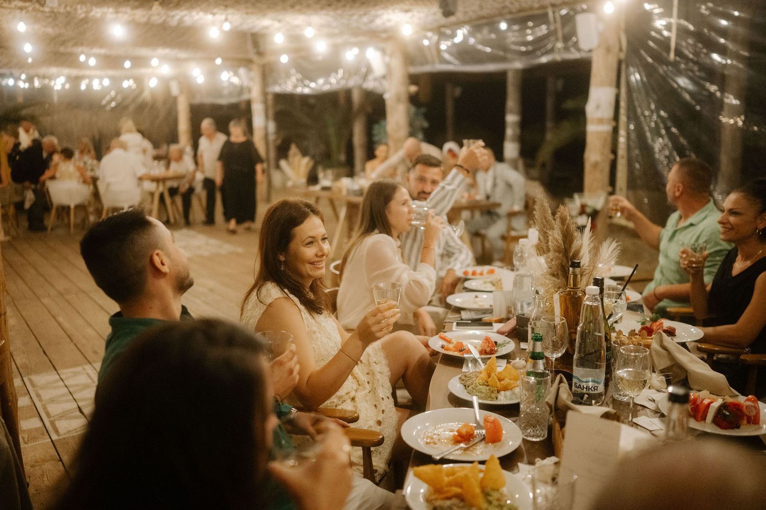 Wedding guests enjoy a festive outdoor dinner reception under glowing string lights on a rustic wooden deck.