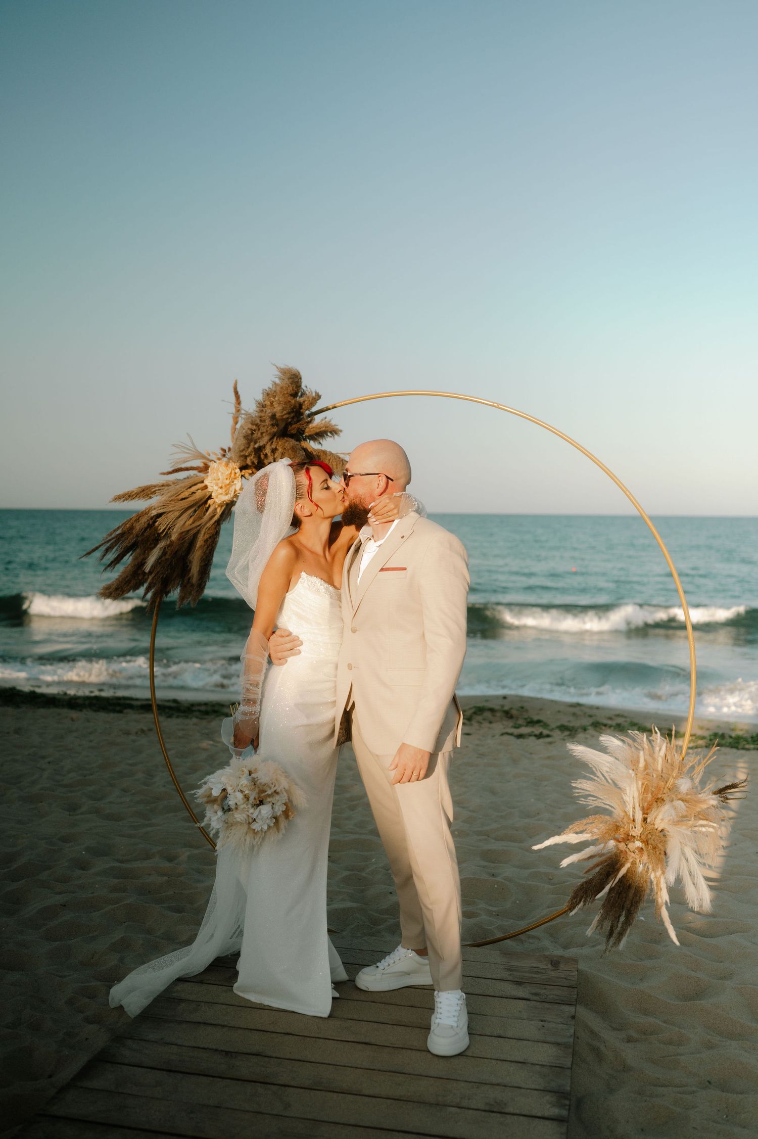 A romantic beachside wedding moment captured under a golden hoop arch adorned with pampas grass.