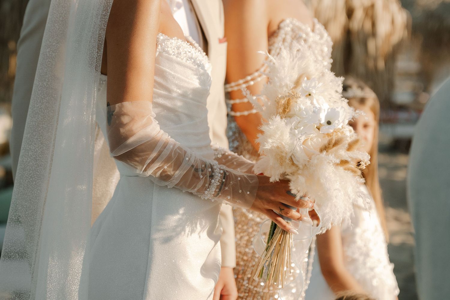 Close-up detail of elegant bridal gown with delicate lace sleeves and feathered bouquet in warm golden light.