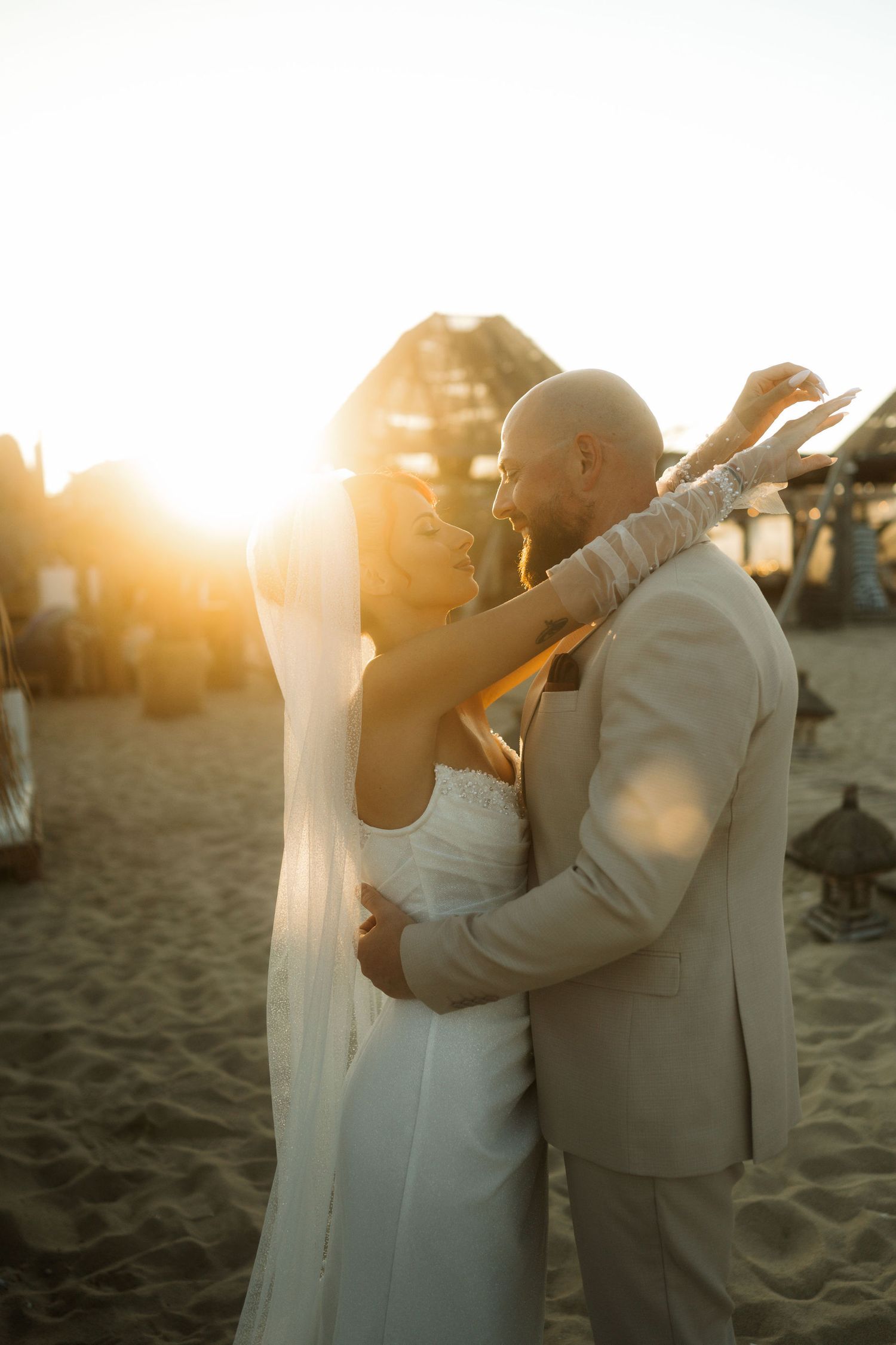 A romantic beach wedding couple embraces during golden hour sunset, with the bride in white and groom in beige suit.