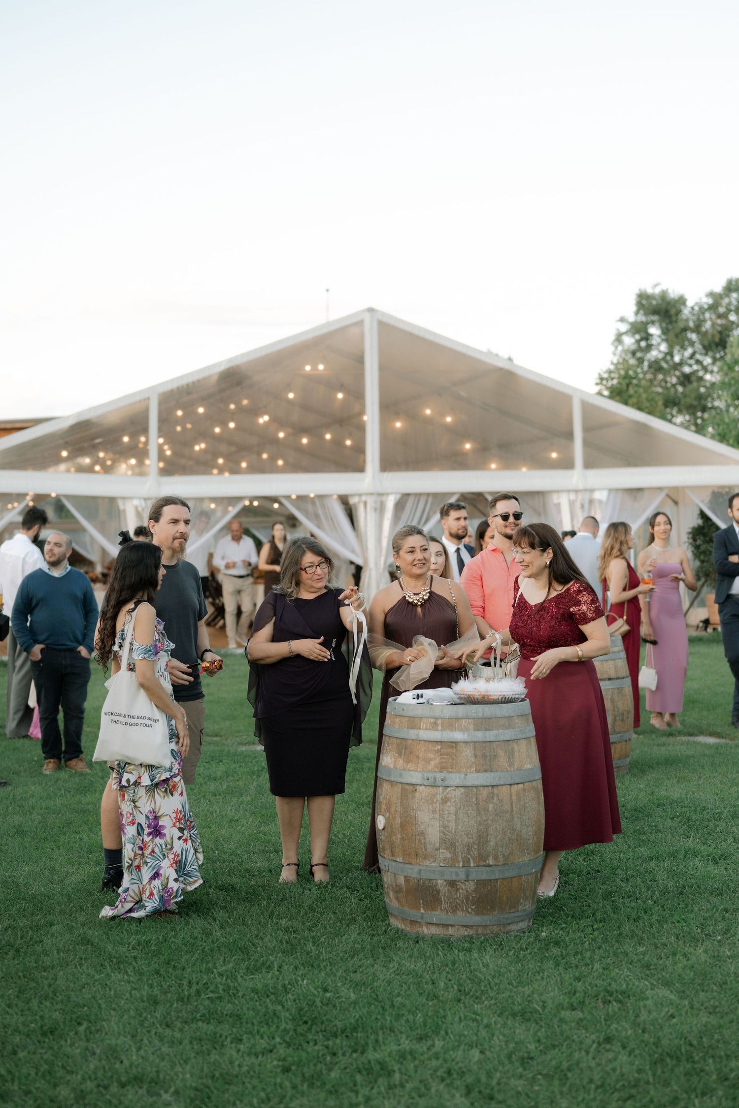 Wedding guests gather around rustic wooden barrels on a grassy lawn under a white tent with string lights.