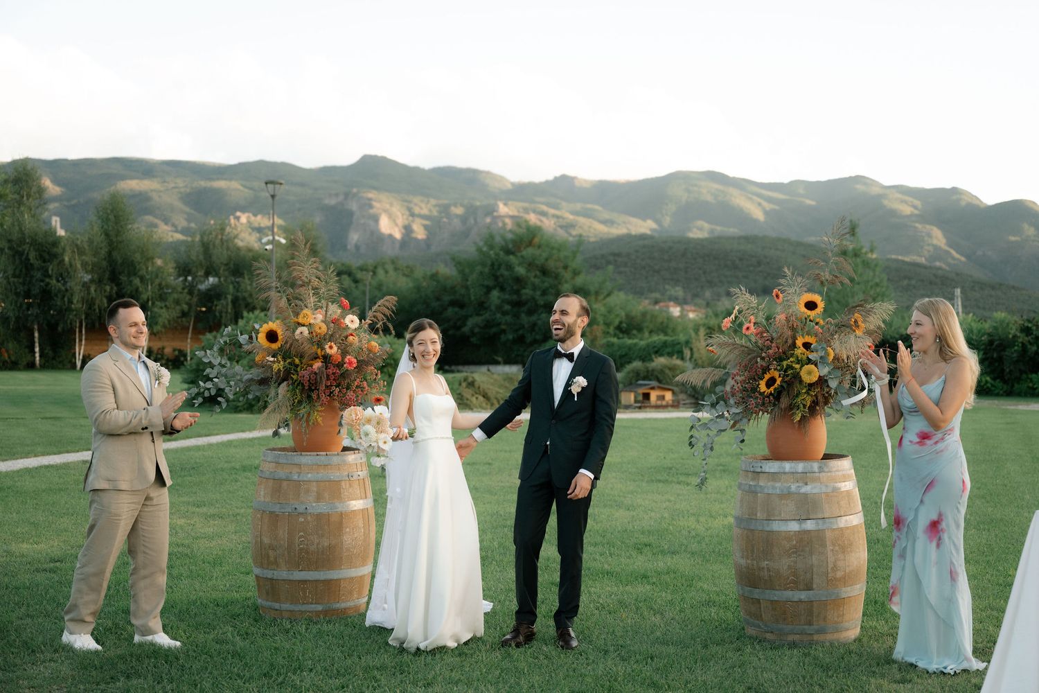 Wedding party gathered around rustic wine barrels during an outdoor mountain ceremony at sunset.