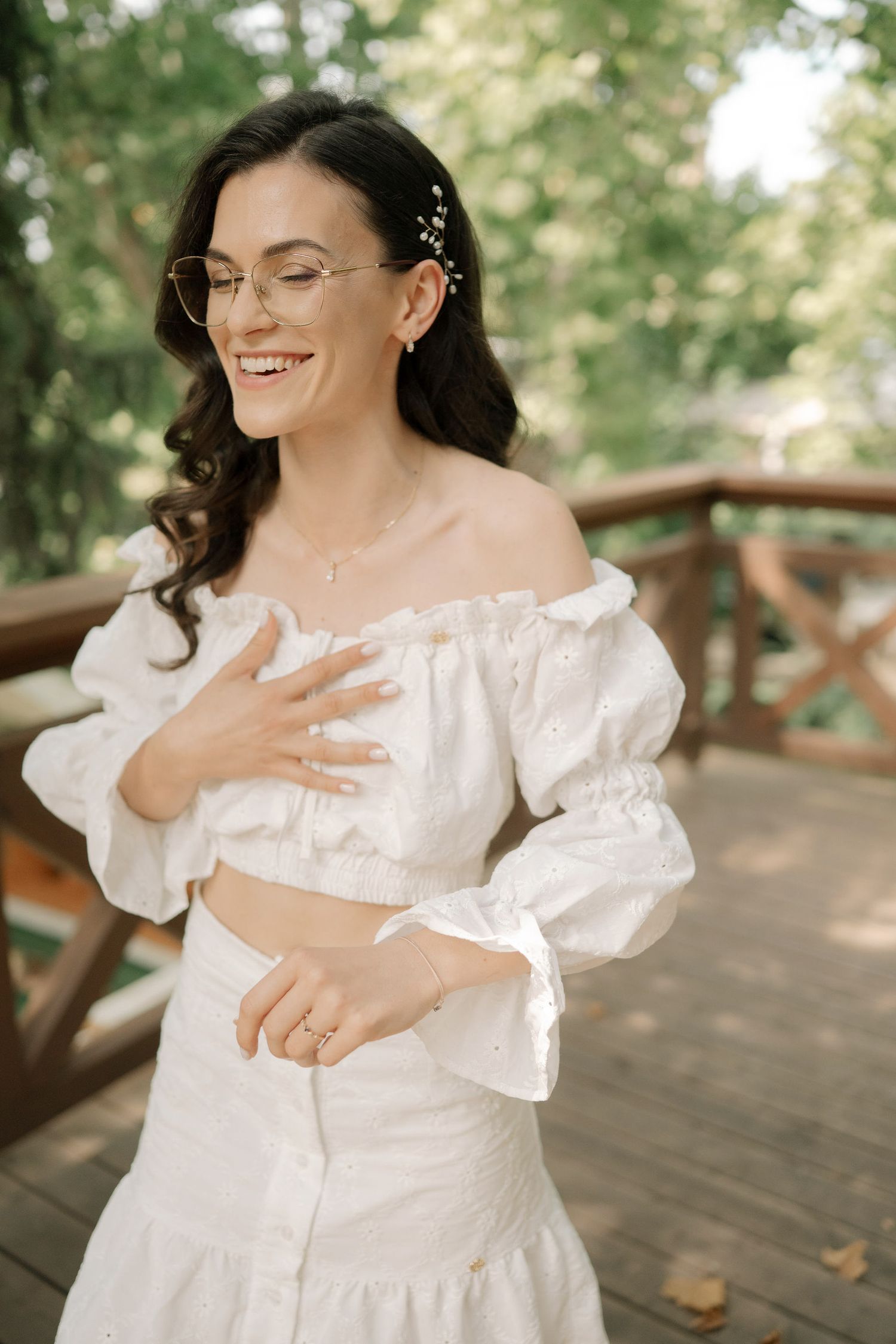 A person in a white dress with puff sleeves stands on a wooden deck surrounded by greenery in a natural setting.