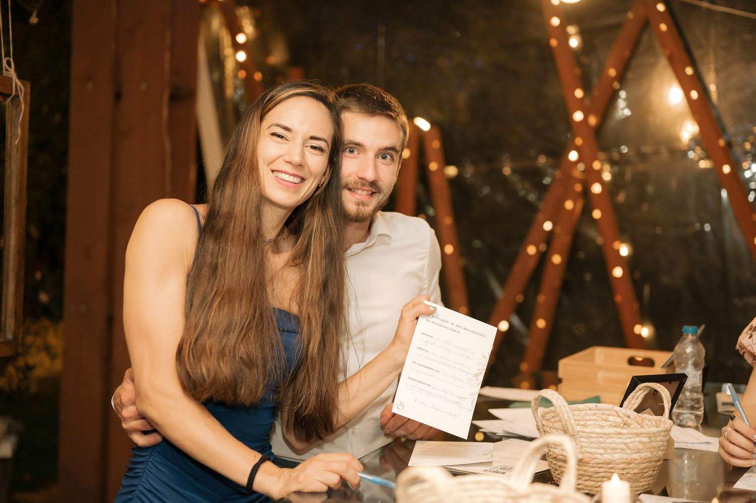 A couple embraces and smiles while holding menus at a cozy restaurant with warm lighting and decorative elements.
