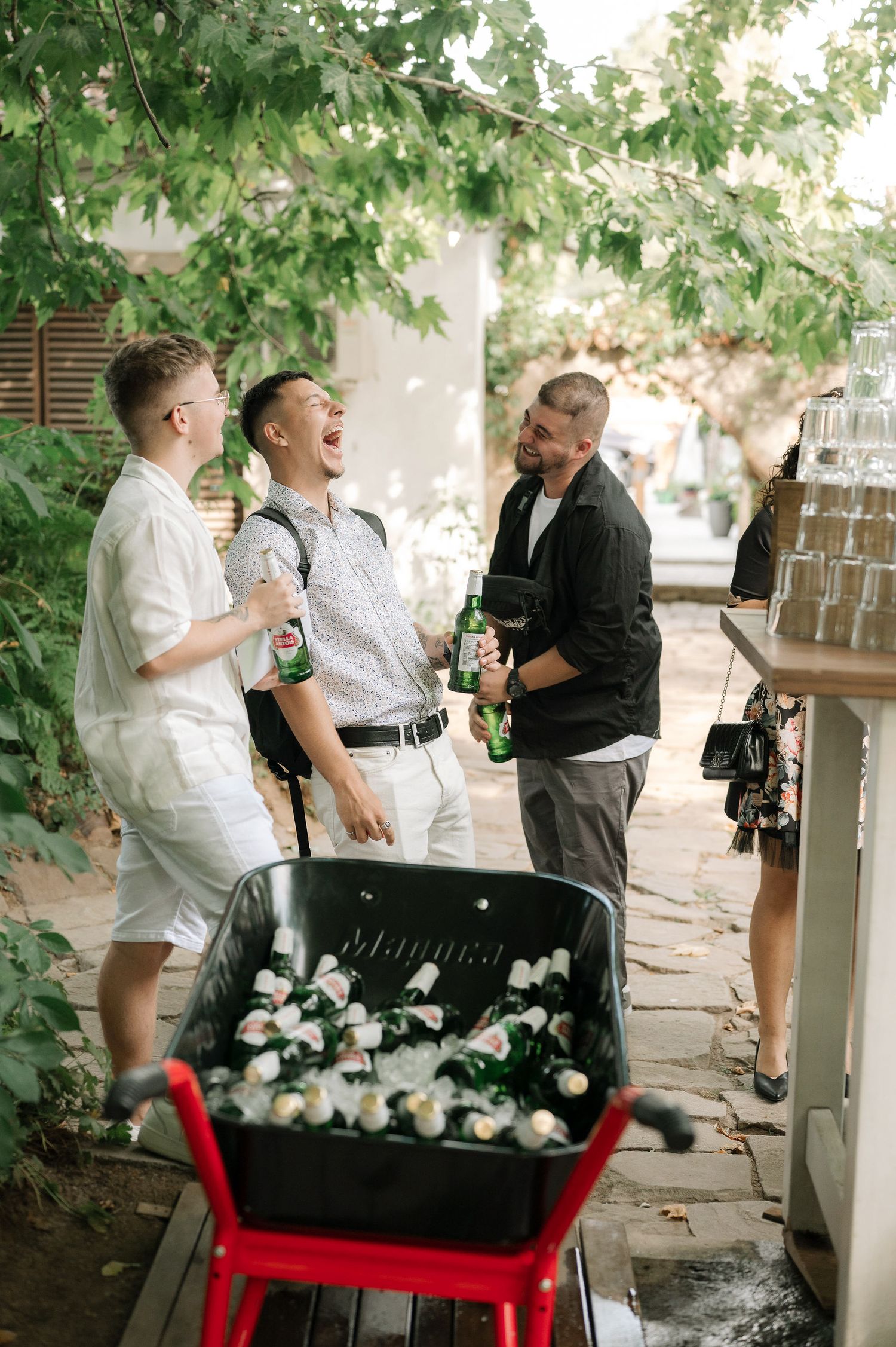 A group of people share joyful moments in a garden setting with a red bench and overhanging green foliage.