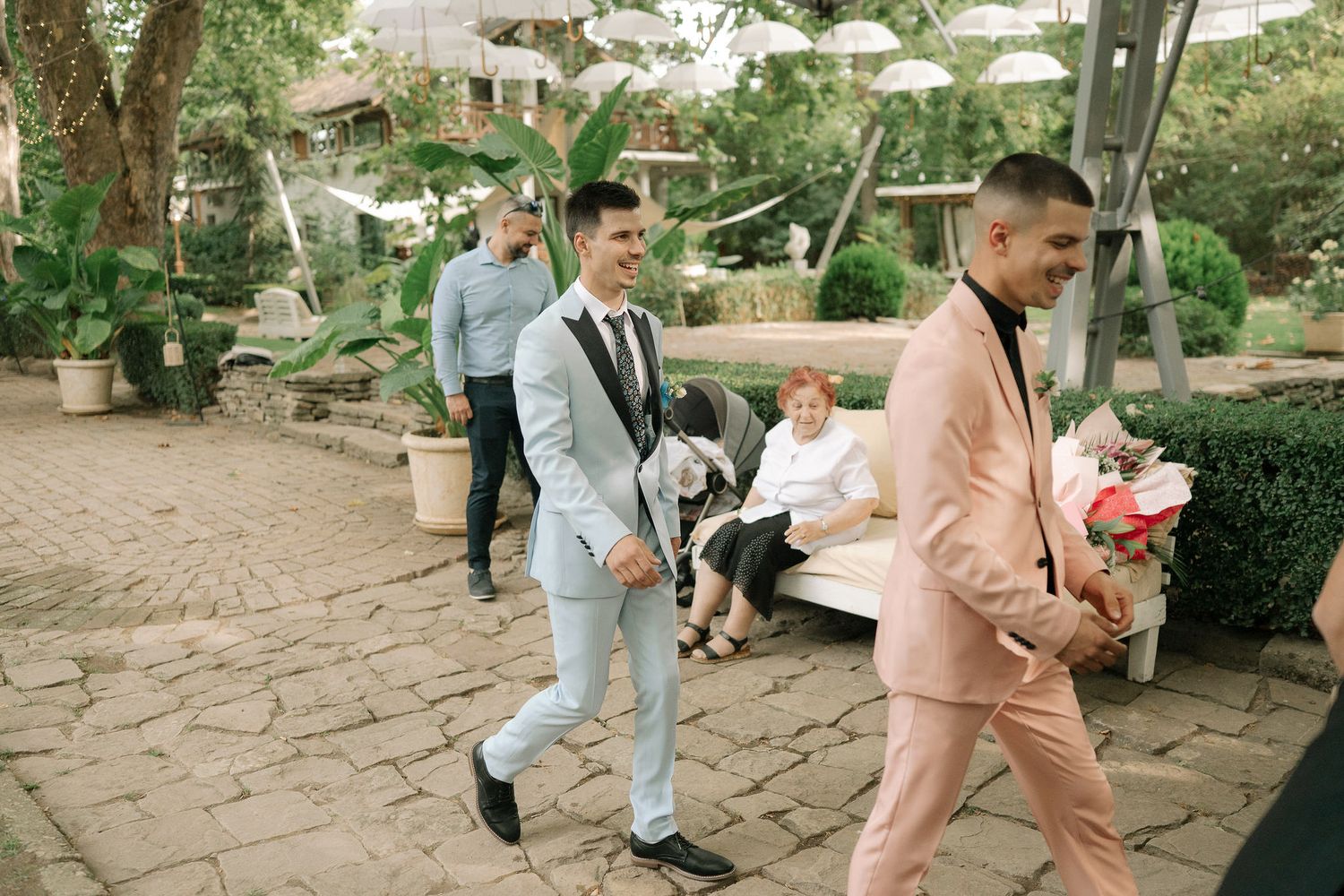 People walking and sitting in a stone-paved garden or courtyard area with lush greenery and natural surroundings.